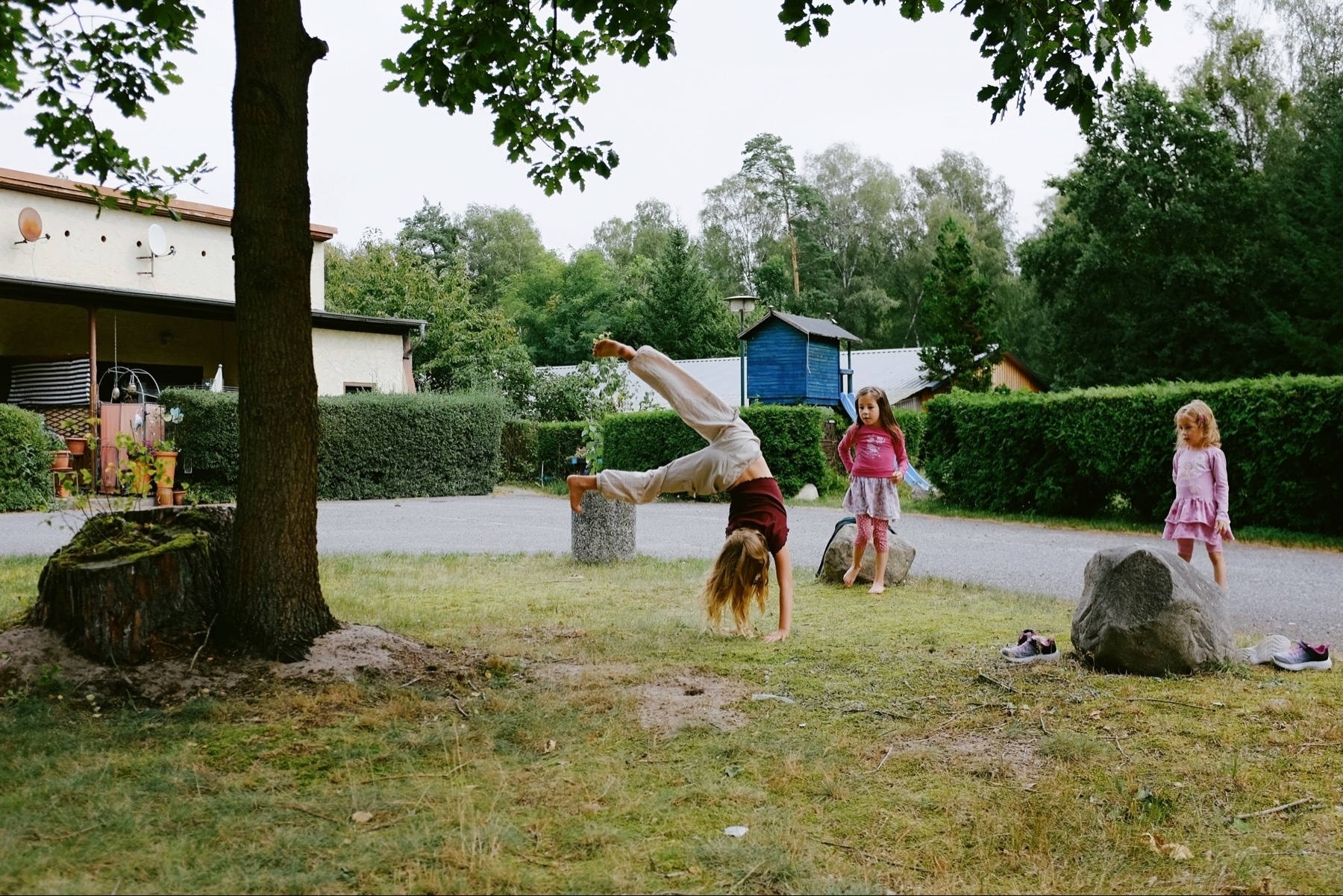 Child doing a handstand on grass with two others watching near a tree, shoes scattered, and a house in the background.