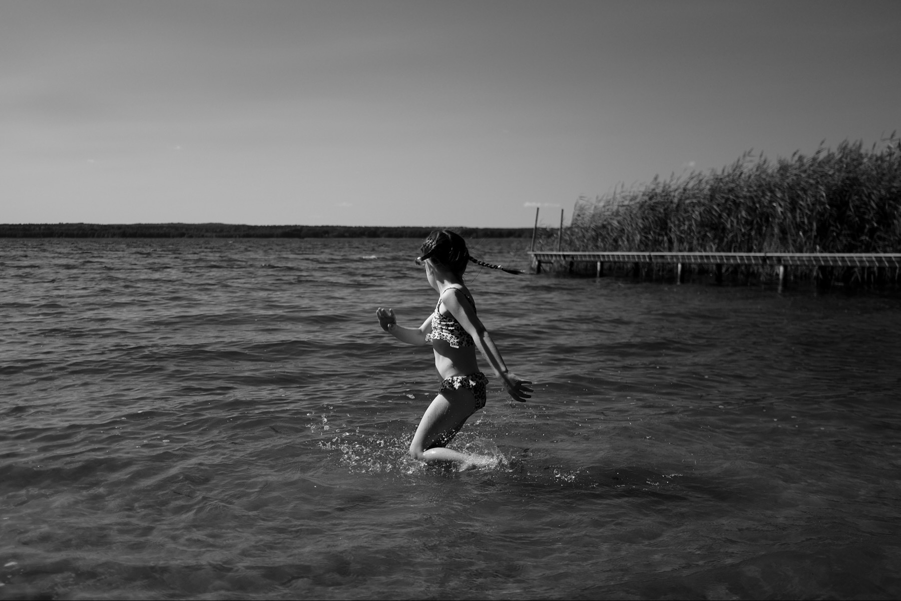 Child dancing on the water at a lakeside, splashing, with reeds in the background.