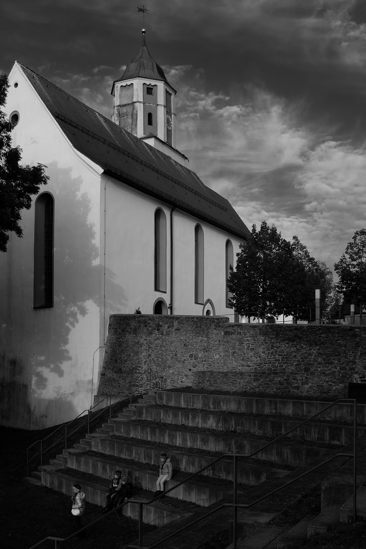 A black and white photograph of a church with a tall, pointed tower. In the foreground, there are stone steps with three children sitting and standing. Trees frame the scene under a cloudy sky.