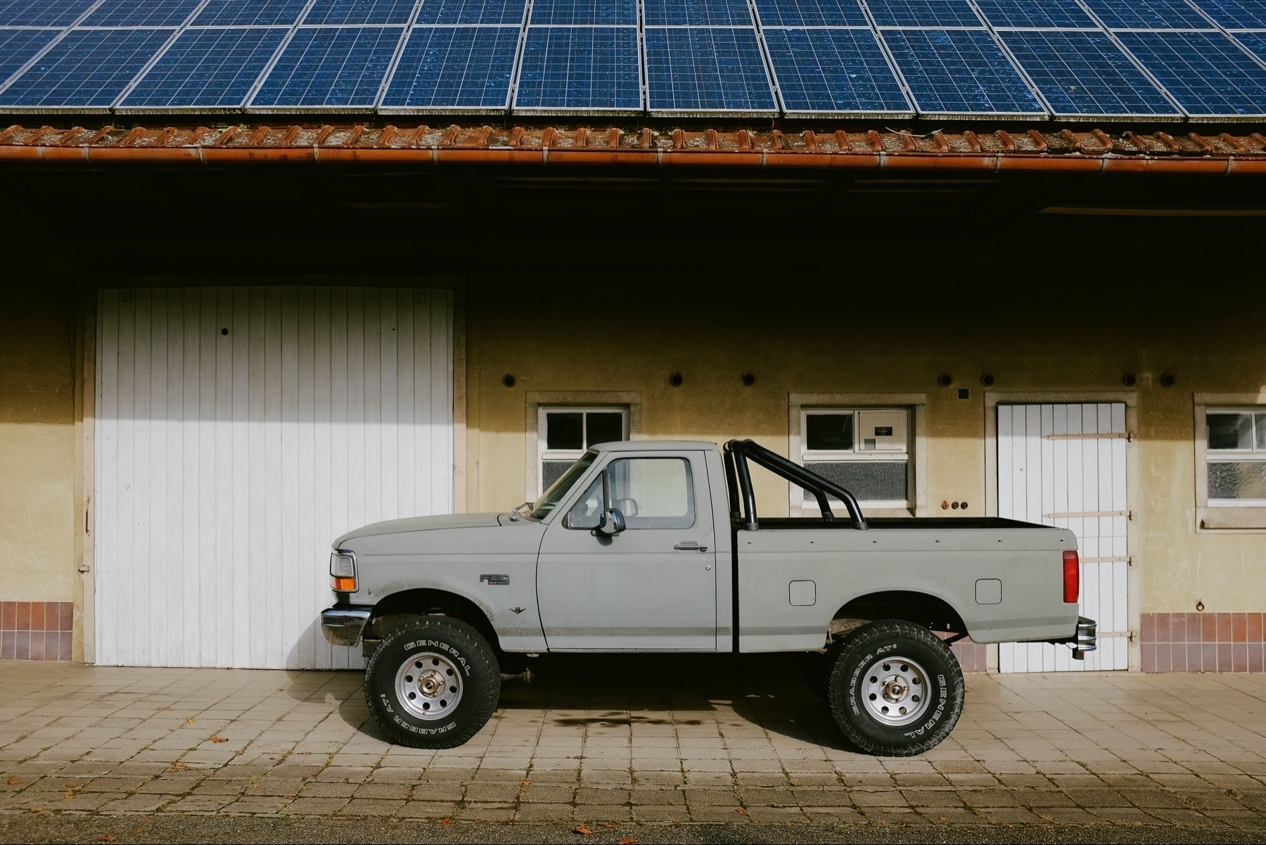 A vintage Ford F150 pickup truck is parked in front of a building with white doors and small windows. The building’s roof is covered with solar panels.