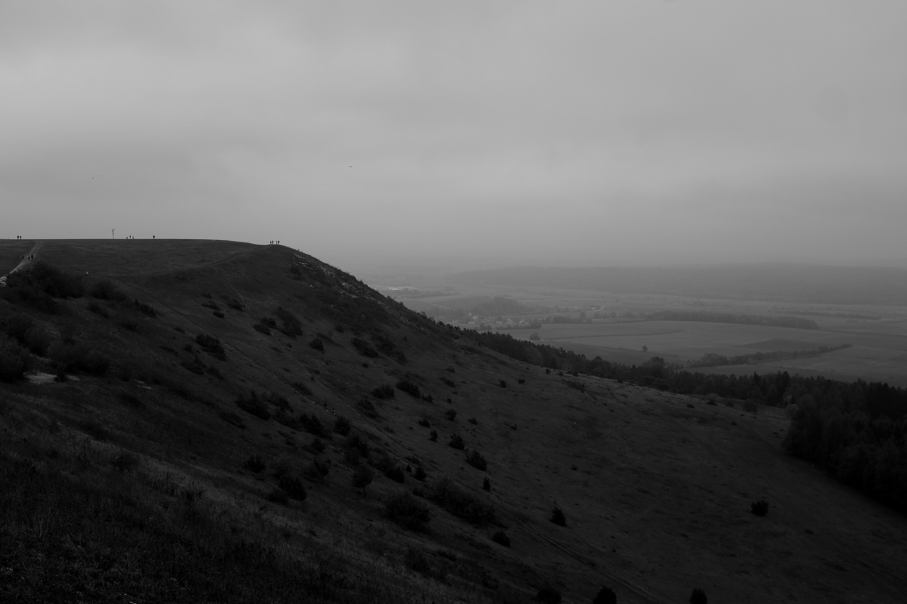 A black and white image of a hilly landscape under an overcast sky. The slope is covered with sparse vegetation, and silhouettes of people are visible on a ridge in the distance. Fields and trees stretch out toward the horizon.