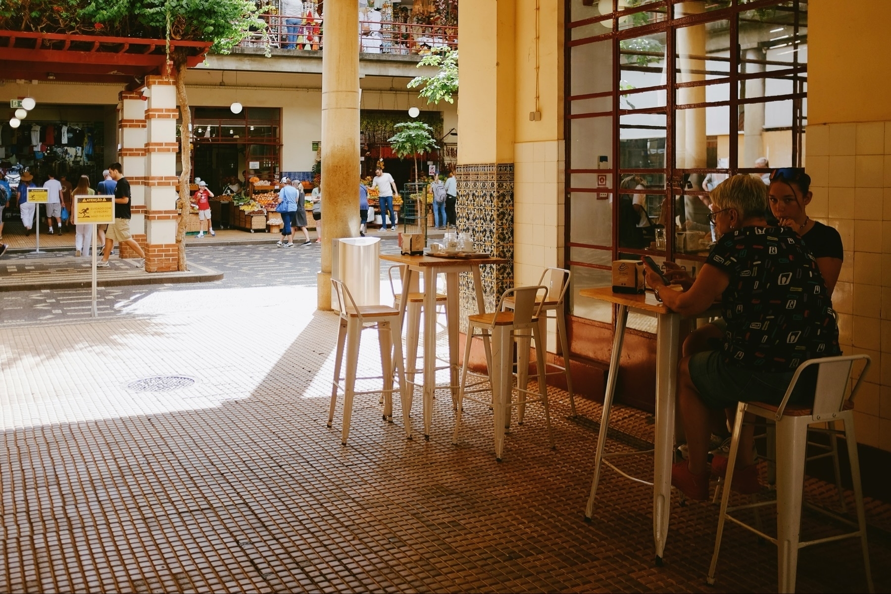 People sitting at a small outdoor café with high stools and tables. In the background, a bustling market scene with people walking near fruit stands and shops. The area is sunlit, with tiled flooring and decorative columns.