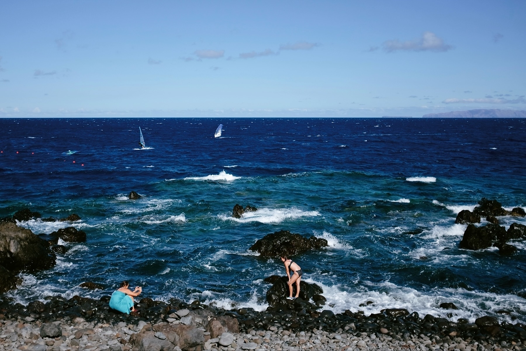 A vibrant image capturing the rocky shoreline of Madeira with the deep blue Atlantic Ocean in the background. Several sailboats are visible on the horizon, dotting the seascape. In the foreground, two individuals are seen sitting on large pebbles near the water’s edge; one is wearing a blue shirt and appears to be using a camera, while the other, in black attire, seems to be looking out at sea. The waves crash against the rocks, creating white foam that contrasts with the ocean’s hue. The sky is clear with few clouds, suggesting a serene day perfect for sailing and photography.