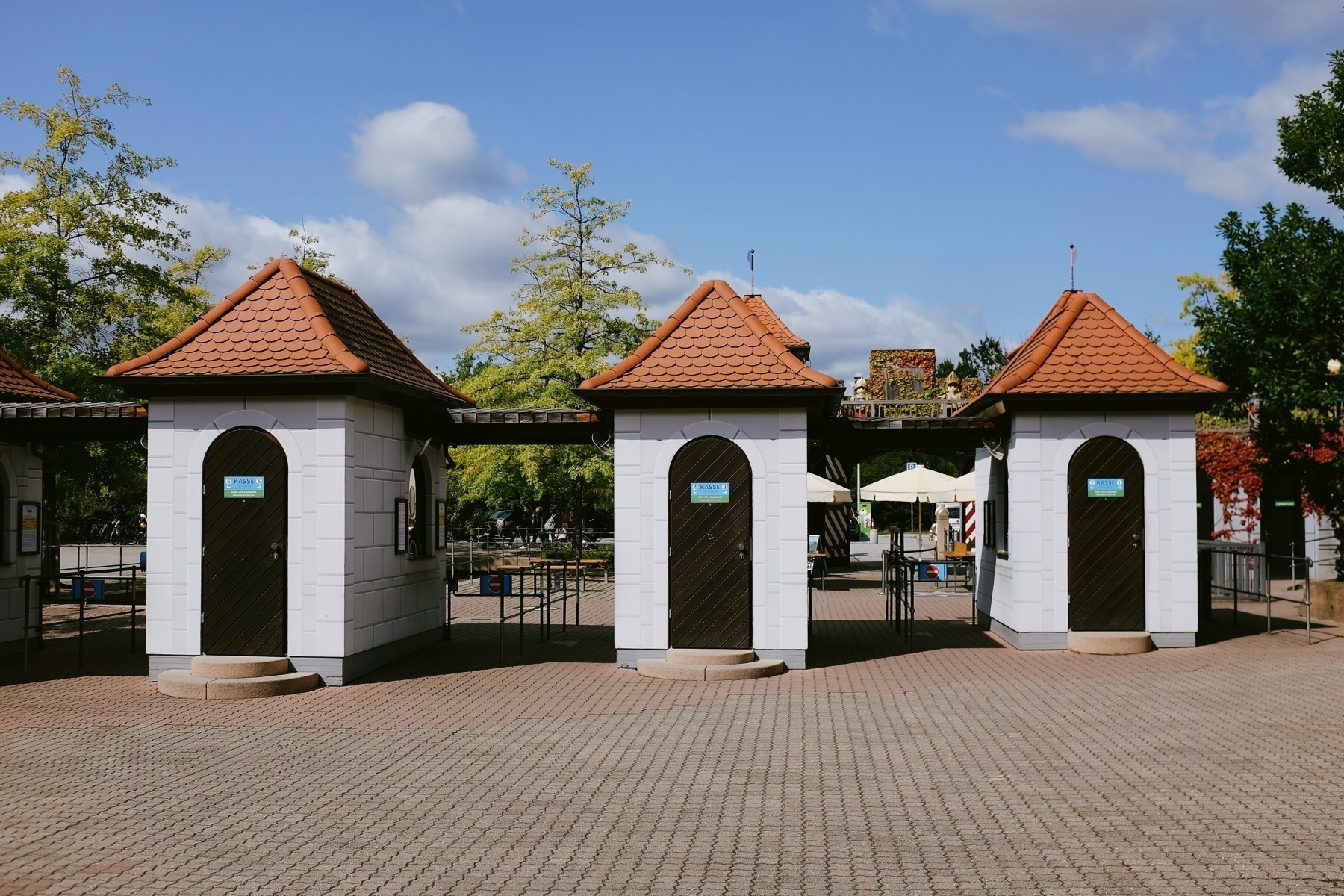 The image shows the entrance gate to an amusement park, featuring three small towers with pointed red tile roofs. The towers are interconnected, with turnstiles and a ticket booth visible between them. Trees and blue sky are in the background.