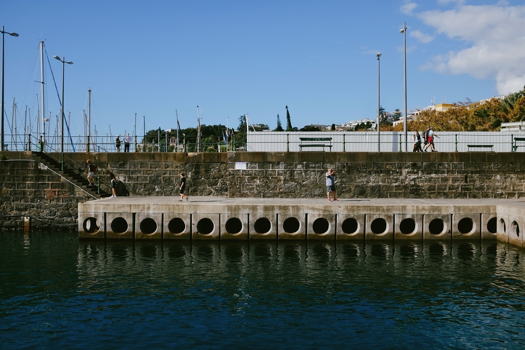 A waterfront scene featuring a concrete dock with circular openings. People are walking and standing on the dock, with boats and masts visible in the background. A clear blue sky and some greenery are also present.