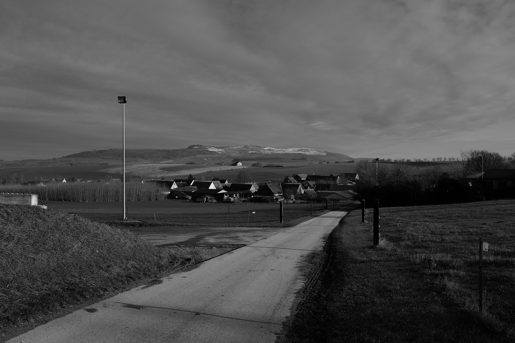 A rural landscape features a narrow road leading towards a small village, set against a backdrop of rolling hills under a cloudy sky.