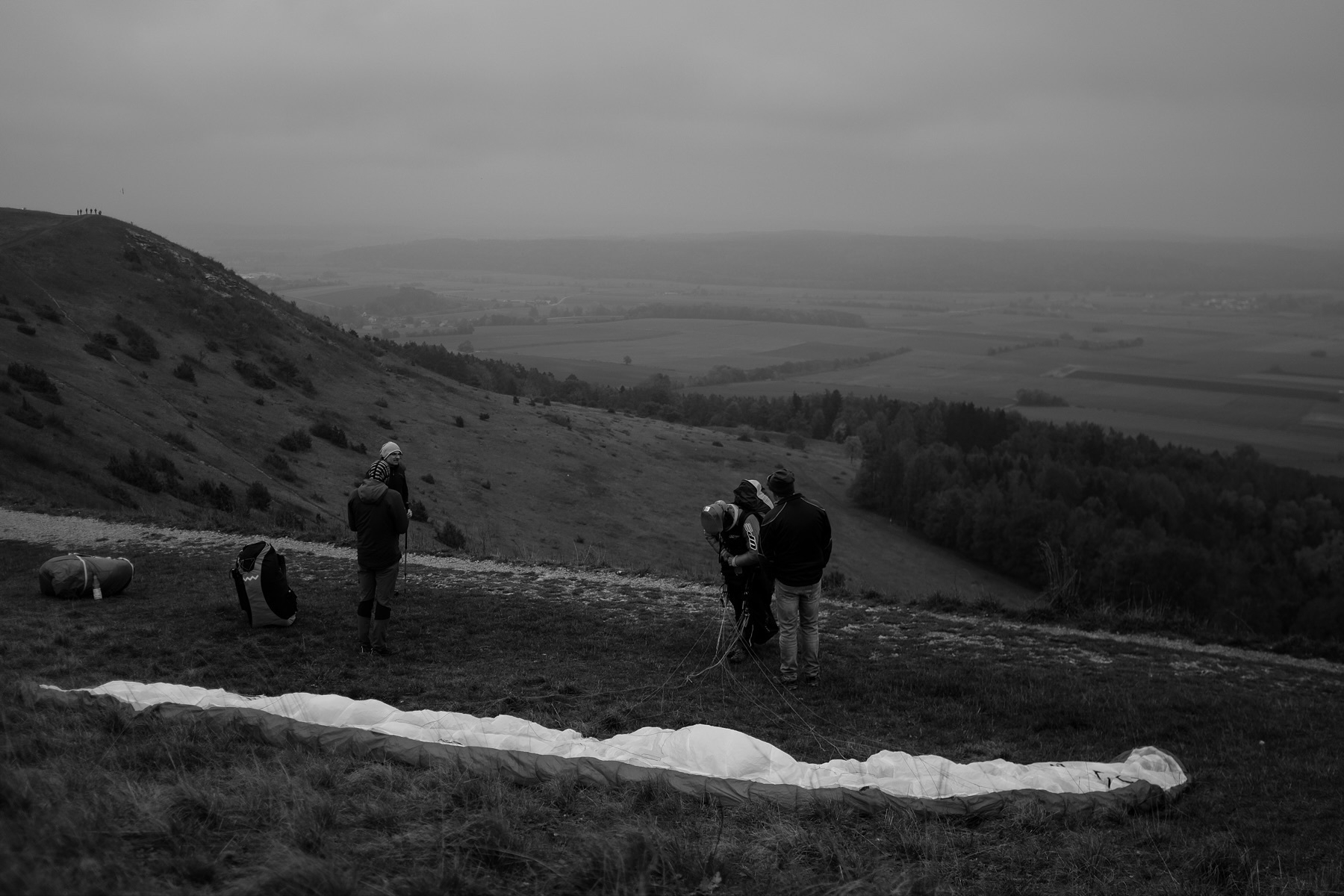 A black and white photo of a hillside where a group of people is preparing a paraglider. The landscape shows rolling hills, fields, and a distant forest under an overcast sky.