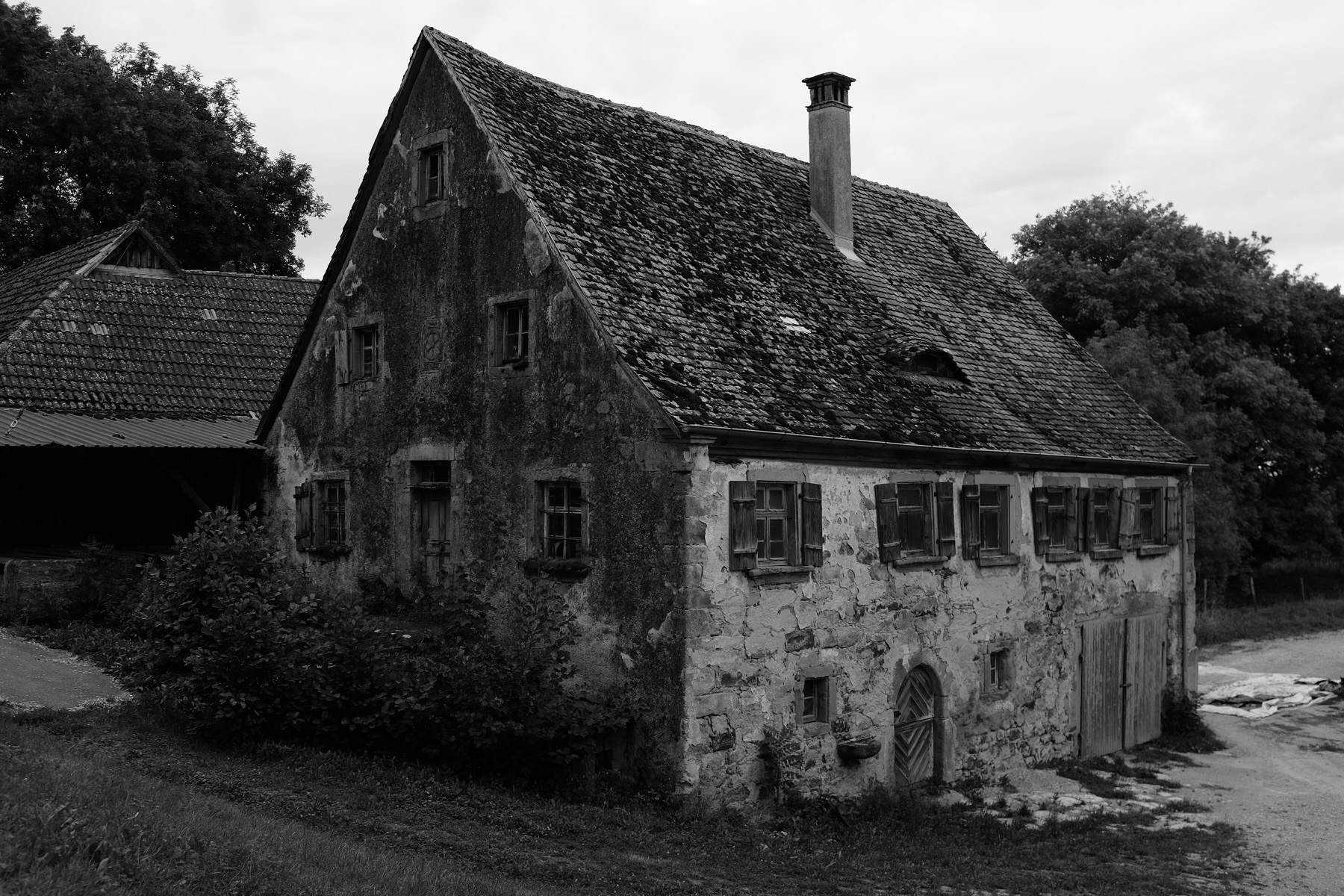 A black-and-white photo of an old, abandoned house with a steep, sloped roof covered in weathered shingles. The building has several small windows with wooden shutters, some of which are broken or missing. The exterior walls are worn and cracked.