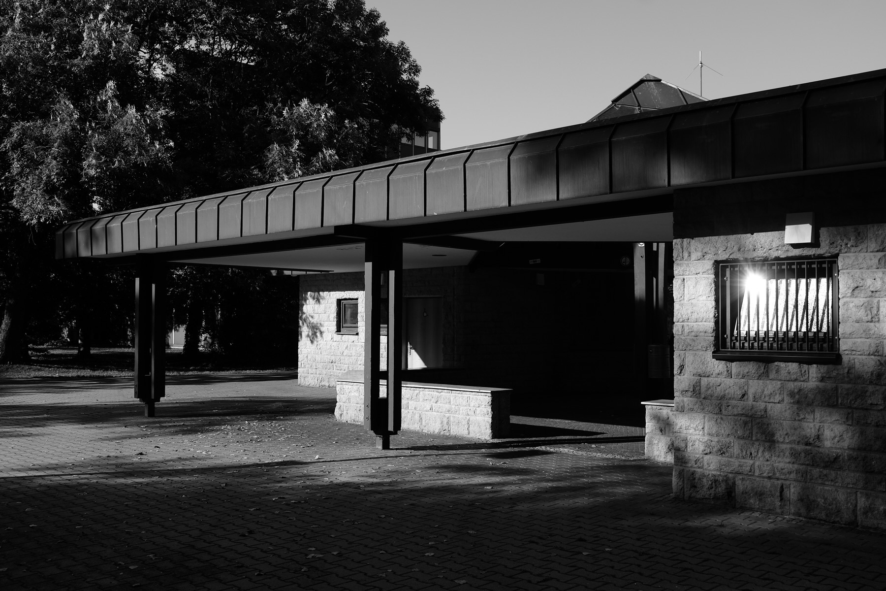 Black and white photograph of a modern building with a prominent overhang and brick facade. There are iron bars on the window and shadows cast by structures and nearby trees. The area is paved with interlocking bricks. The bright sunlight creates stark contrasts.