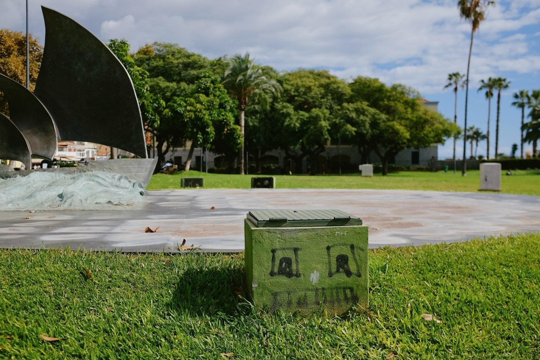 A public park featuring a metal sculpture resembling a sailboat with wave-like designs. In the foreground, a green utility box with black graffiti is visible. The background shows trees, grass, and a partly cloudy sky.