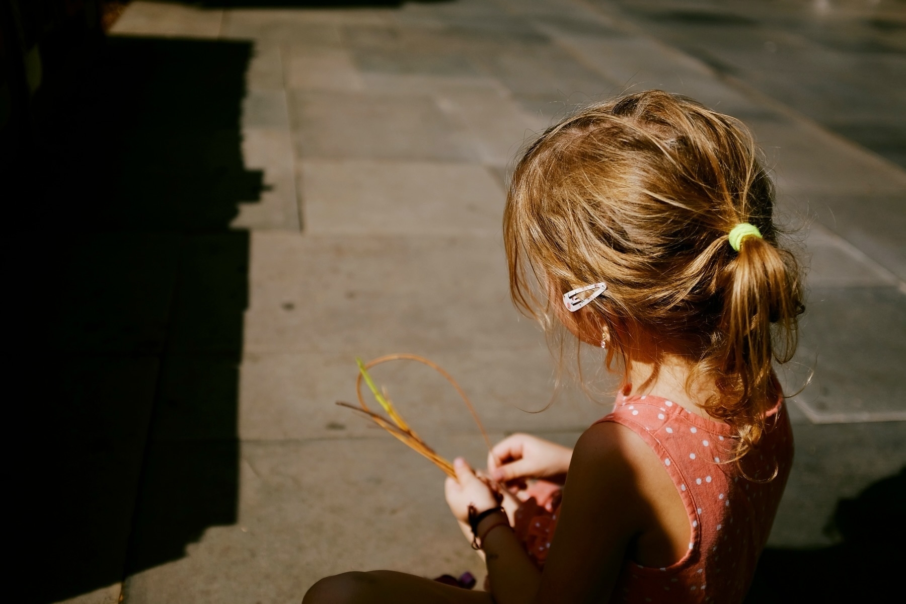 A young girl with a ponytail, secured with a yellow hair tie and a hair clip, sits on the pavement in a pink polka dot dress, playing with a few small sticks. The sunlight casts her in partial shadow.