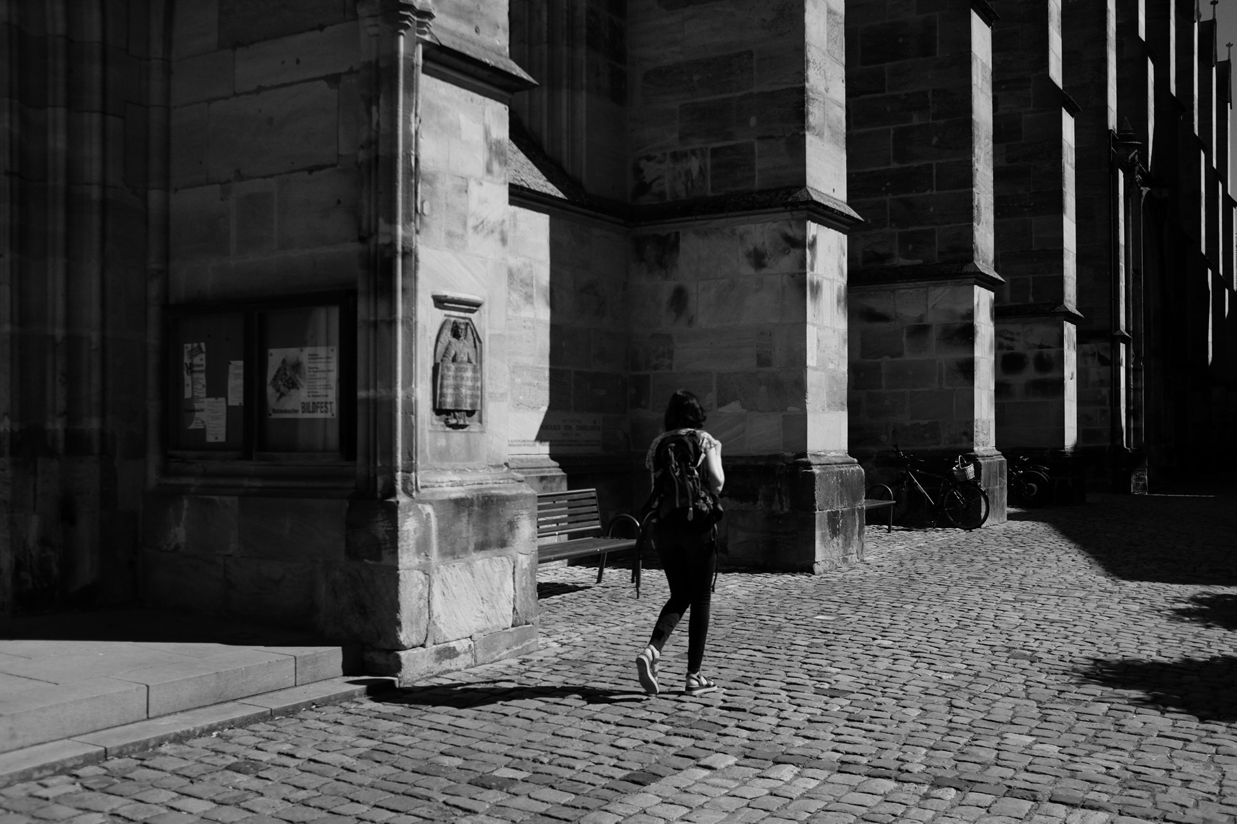 A person wearing a backpack walks on a cobblestone street beside a large stone building. The scene is in black and white, with dramatic shadows cast by the building’s columns and sunlight illuminating parts of the walkway.