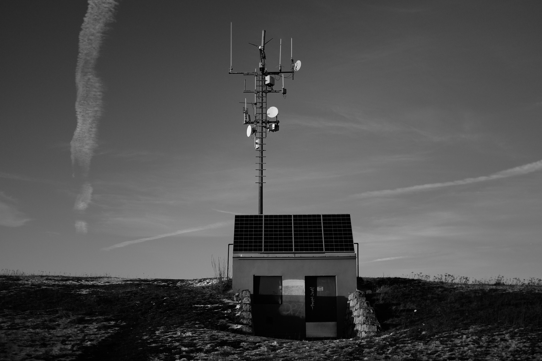 A small, concrete building with solar panels and an antenna tower stands under a sky marked by jet trails.