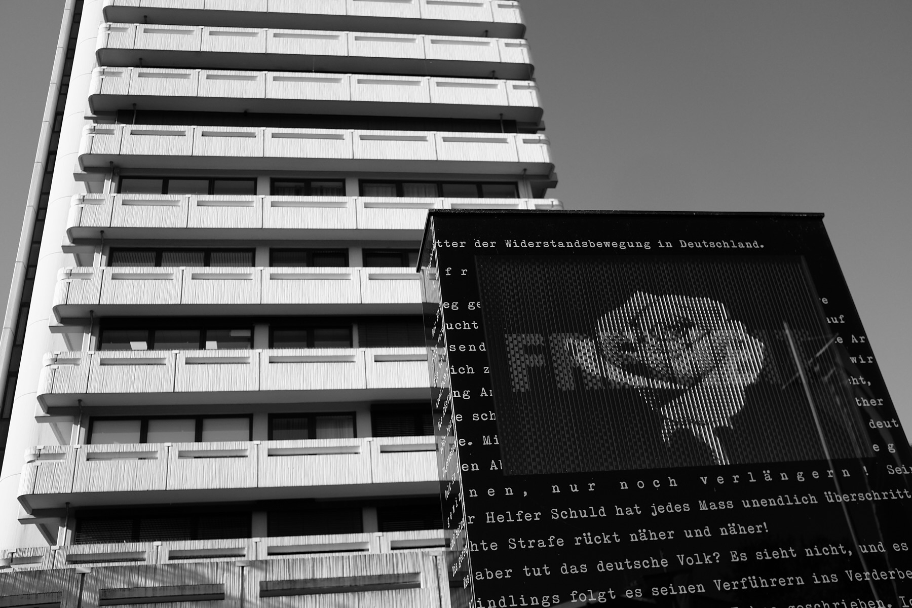 A black and white photograph featuring a modern, multi-story building with rows of balconies. In the foreground, there is a large monument about the White Rose, a non-violent, intellectual resistance group in Nazi Germany.