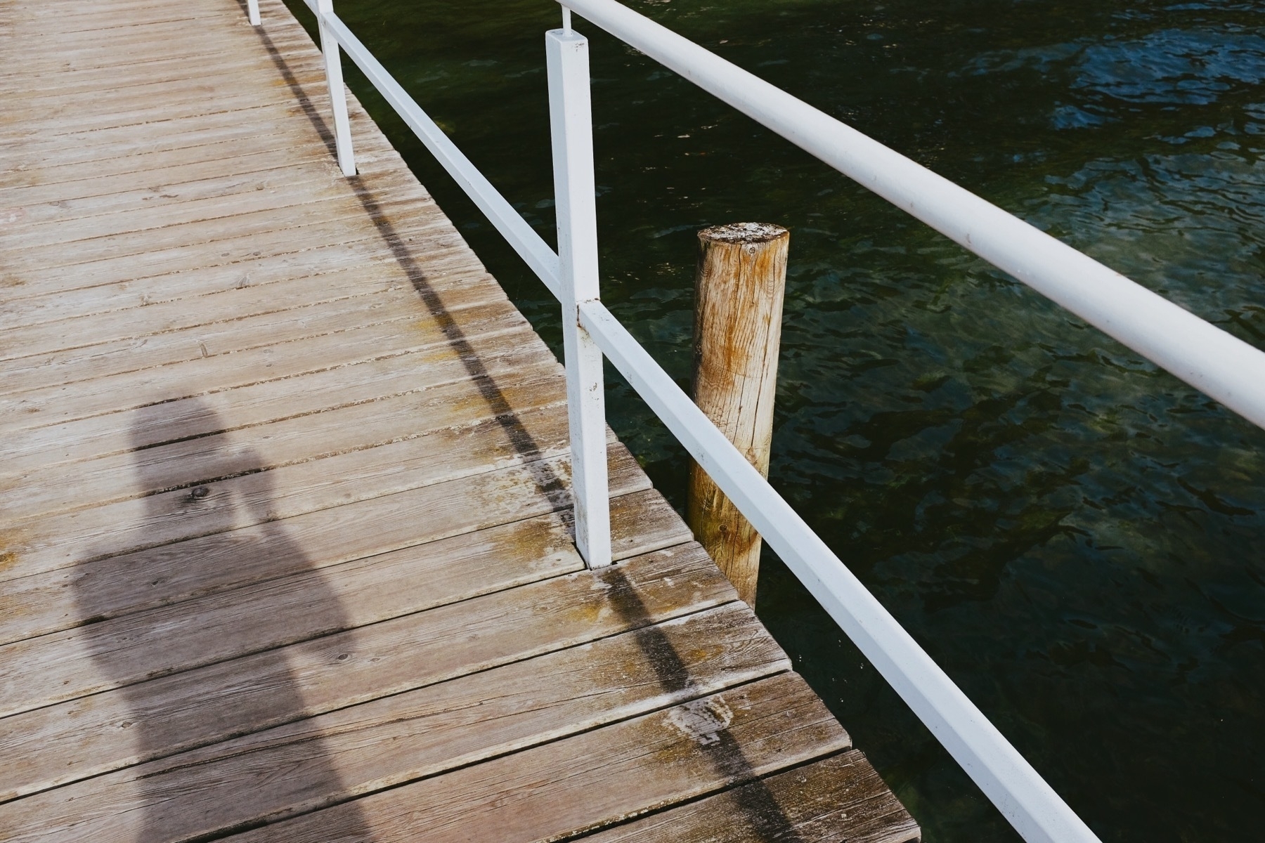 Wooden pier with white railing over water, shadow of a person taking a photo on the deck, and a single mooring post.