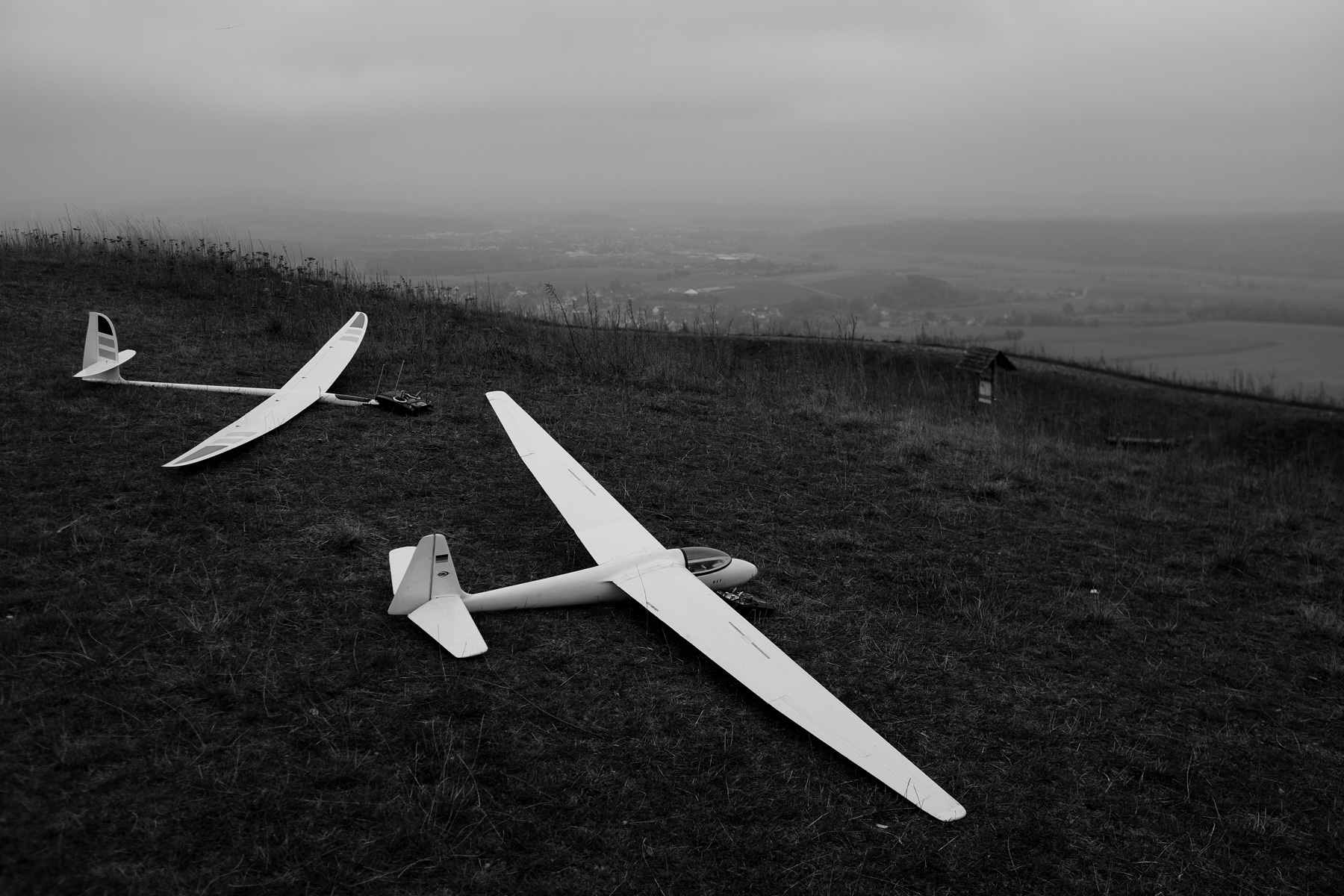 Two white model gliders resting on a grassy hilltop under a cloudy sky, with a distant view of the landscape in the background.