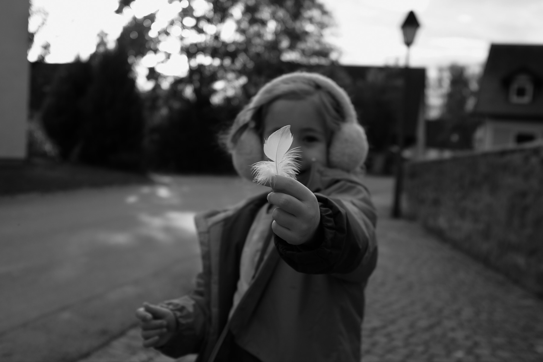 A child wearing earmuffs and a winter coat is holding a feather towards the camera on a cobblestone street. The background is slightly blurred, showing trees and houses. The image is in black and white.