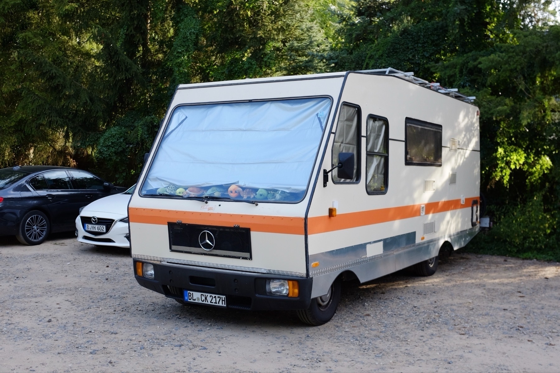 White and orange camper van parked on a gravel lot, surrounded by lush green trees.