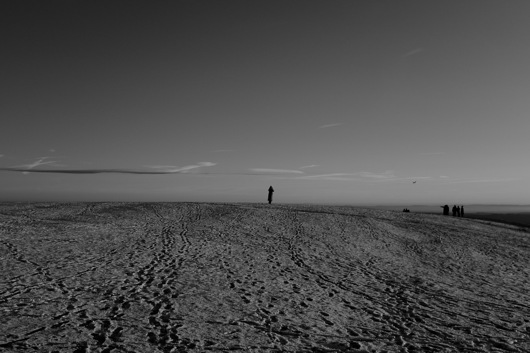 A lone figure stands on a vast, textured landscape beneath an expansive sky, with a group of people visible in the distance.