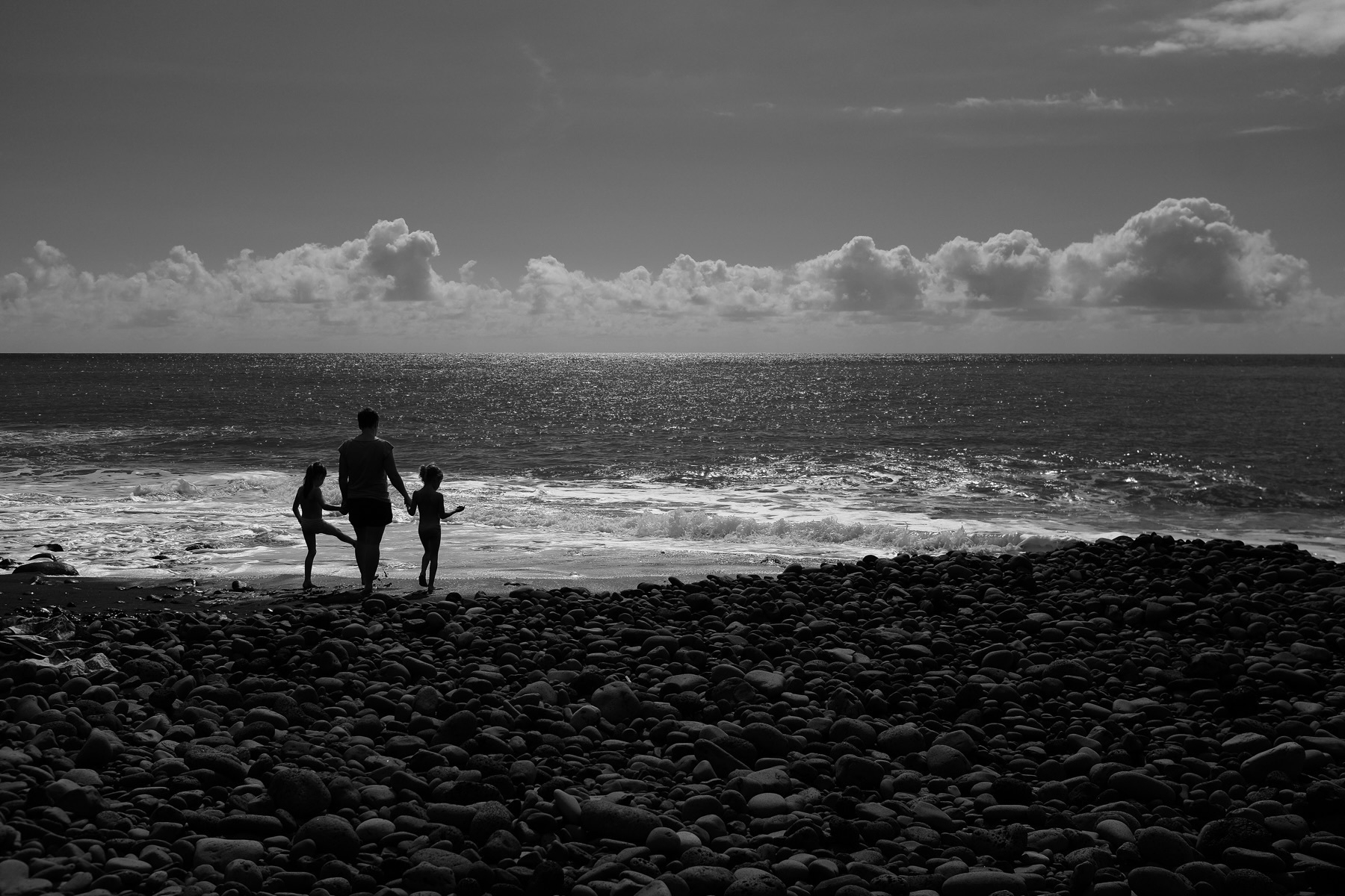 A black and white silhouette of an adult and two children walking towards the ocean on a rocky beach. The sky is partly cloudy, and the sun reflects on the water.