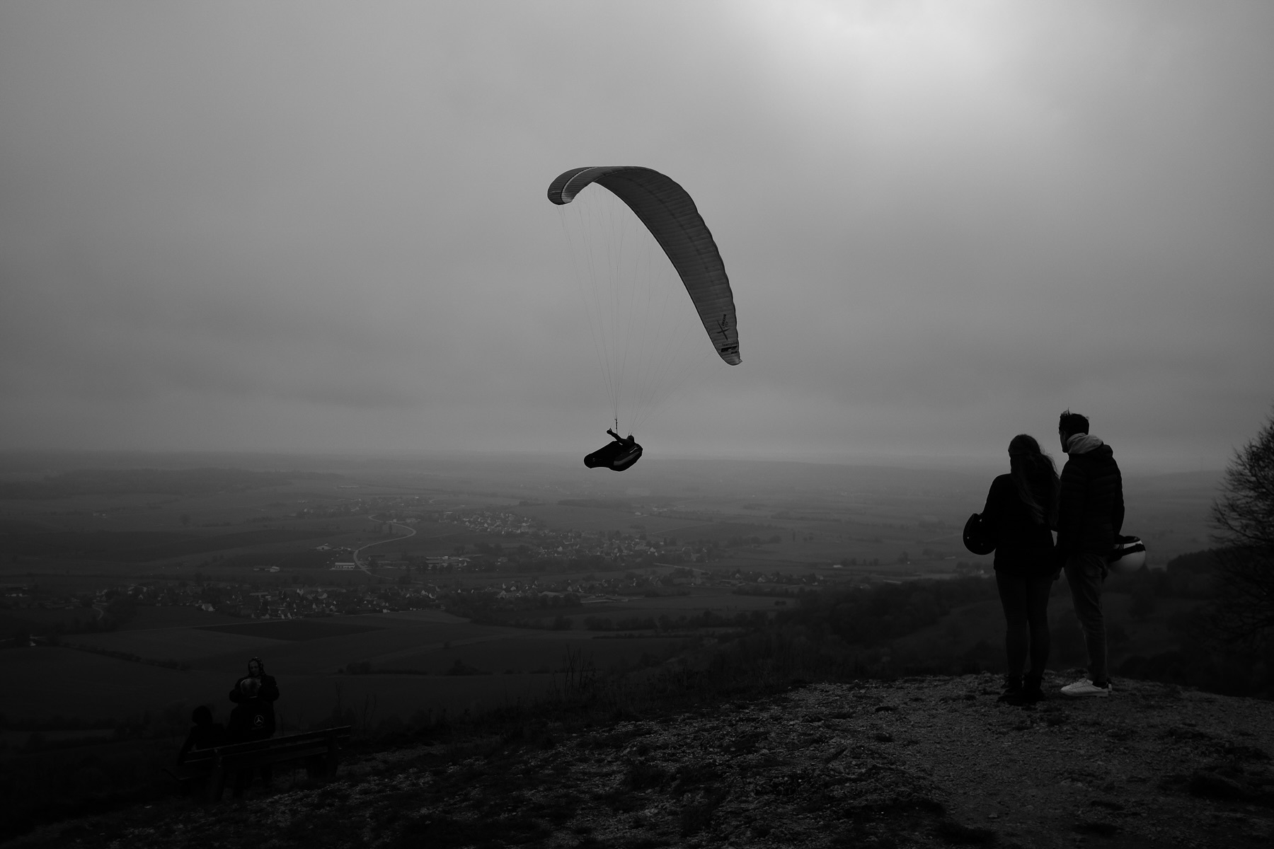 A black and white image of a paraglider soaring above a vast landscape with fields and a town in the background. Two people stand on a hill in the foreground, watching the paraglider. The sky is overcast, creating a moody atmosphere.