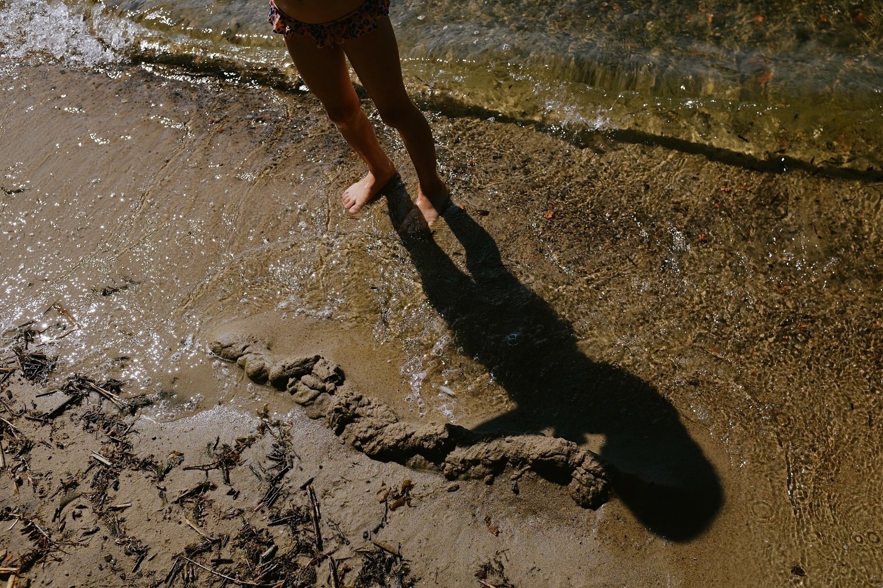 Bare feet by water’s edge with shadow and sandcastle ruins.