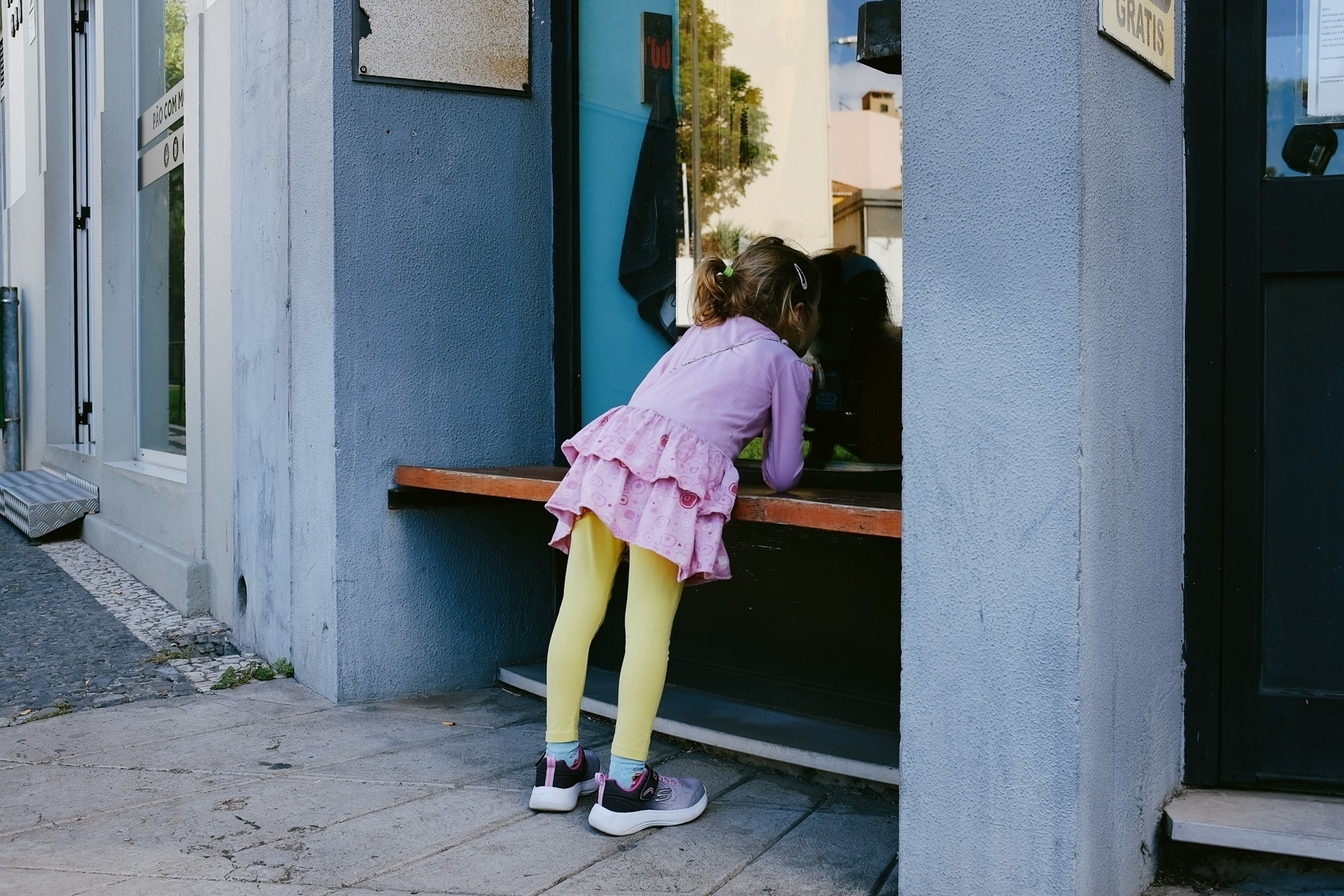 A young girl in a purple top and pink skirt with yellow leggings leans on a ledge, looking inside a window. She is wearing sneakers, and her hair is tied in a ponytail. The scene is on a sidewalk next to a building