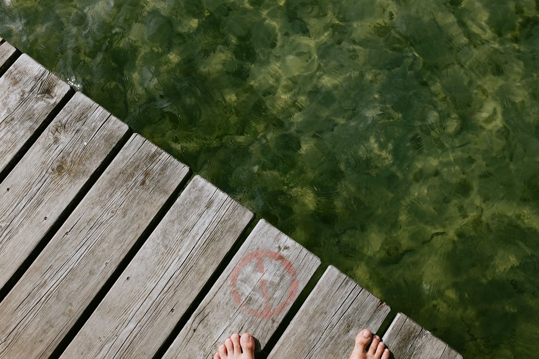 Wooden dock over greenish water with visible rocks below, a person’s bare feet at the edge, and a faded red symbol on one plank.