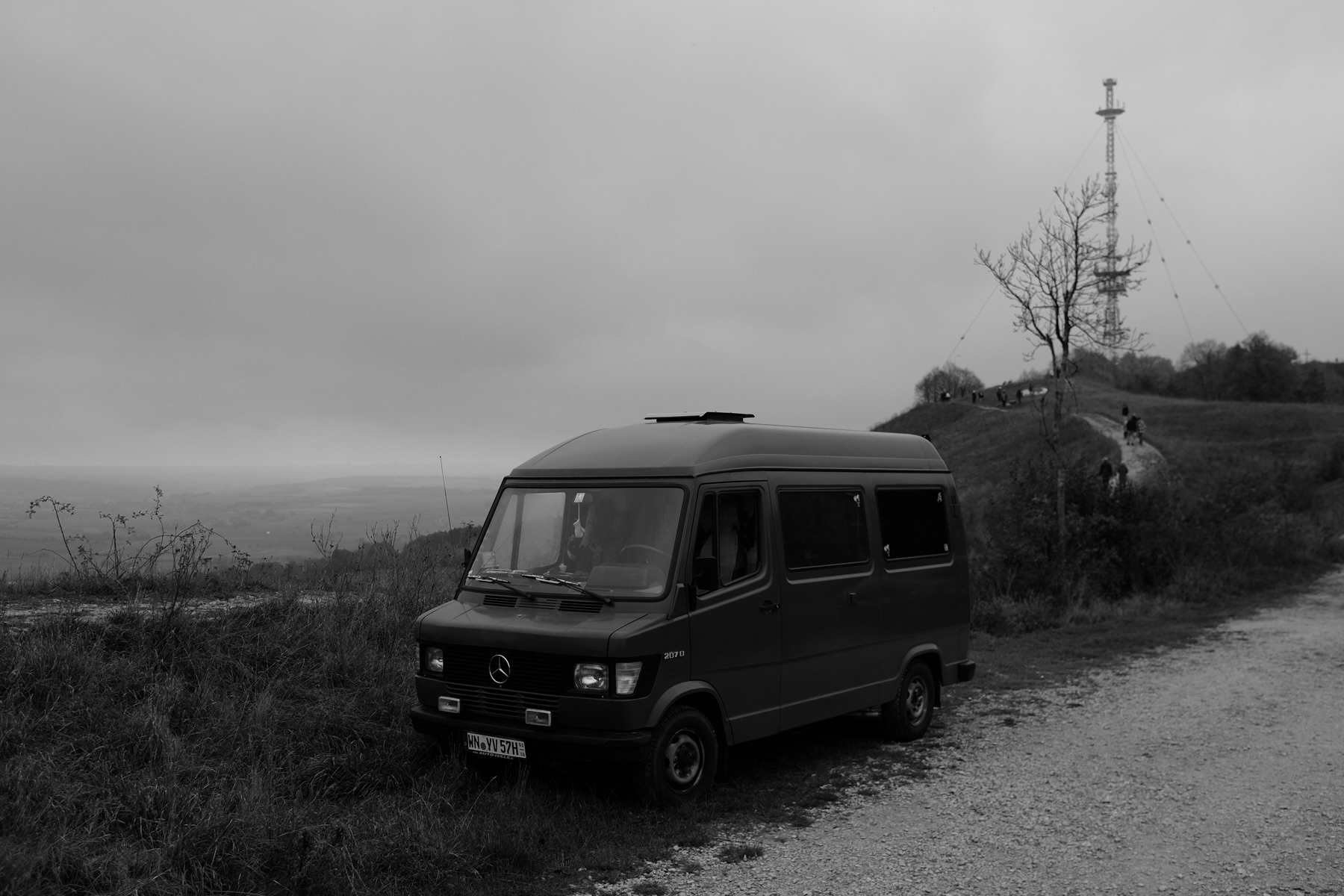 A black and white photo of a Mercedes-Benz van parked on a gravel path next to grassy terrain. In the background, there is a large transmission tower and a small group of people walking along a path. The sky is overcast.