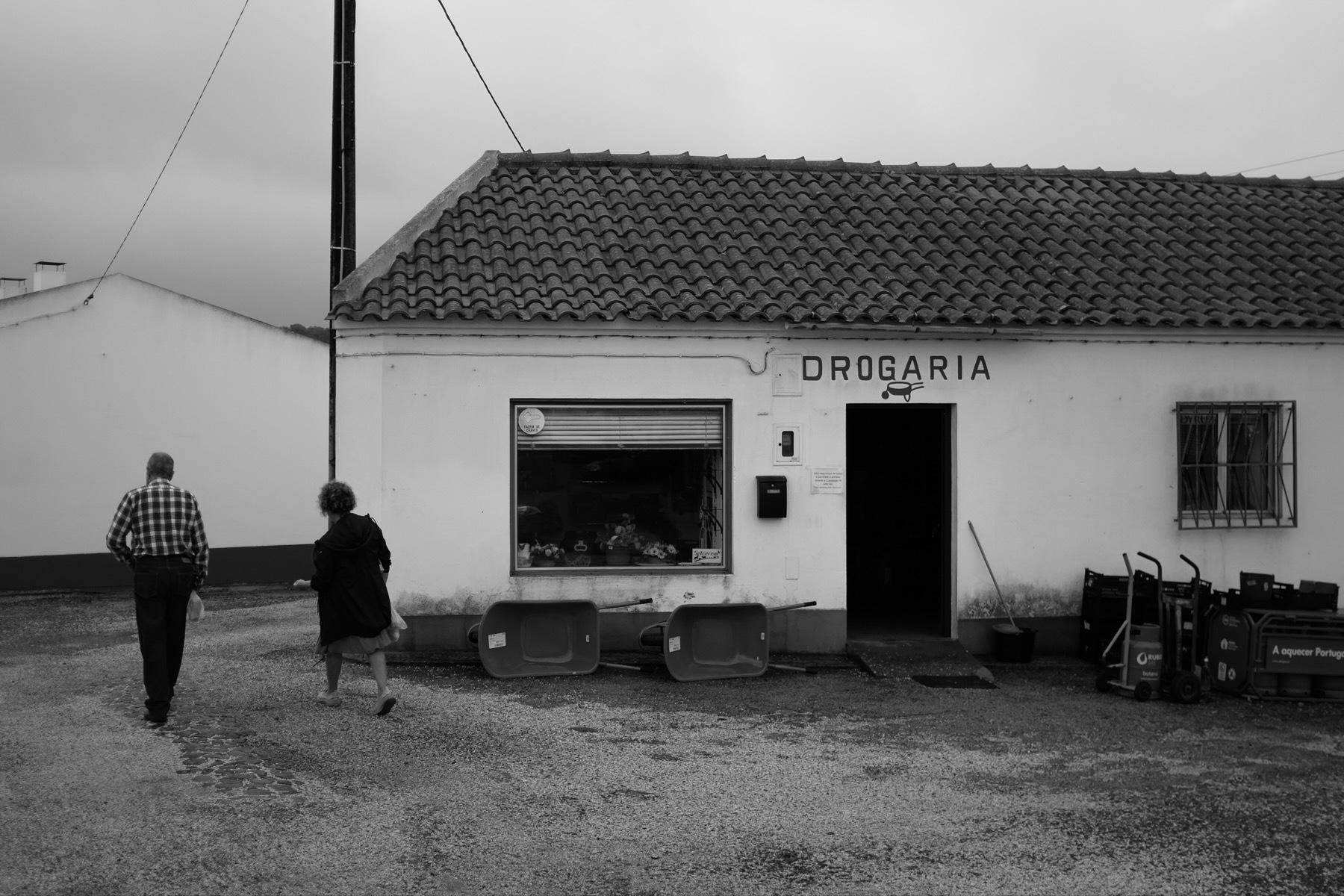 Monochrome photo of two individuals walking past a small shop labeled ‘DROGARIA’ with various items displayed outside.