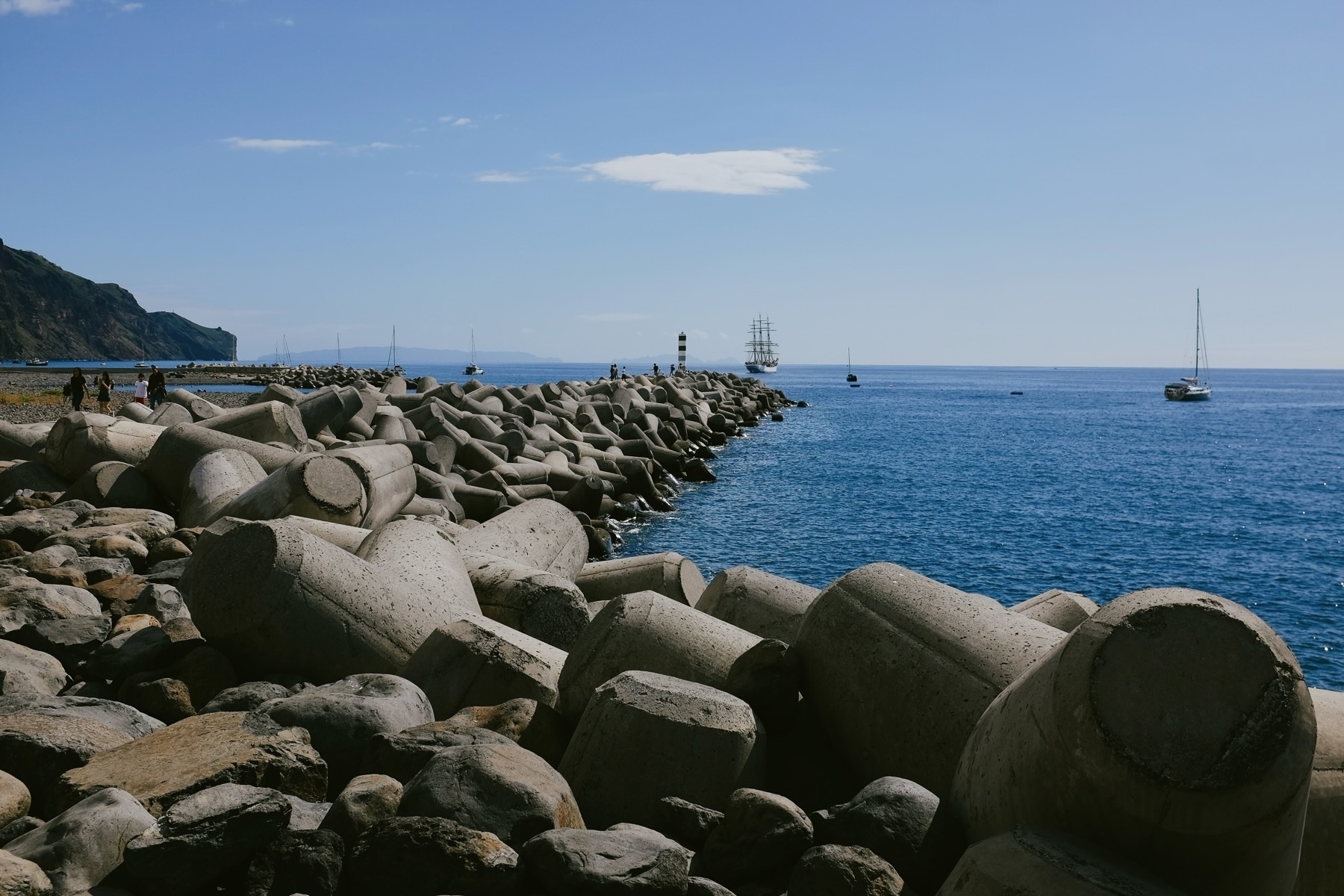 A seaside view featuring large concrete tetrapods forming a breakwater, with people walking along it. In the background, a sailing ship, a striped lighthouse, and several sailboats rest on the calm blue sea.