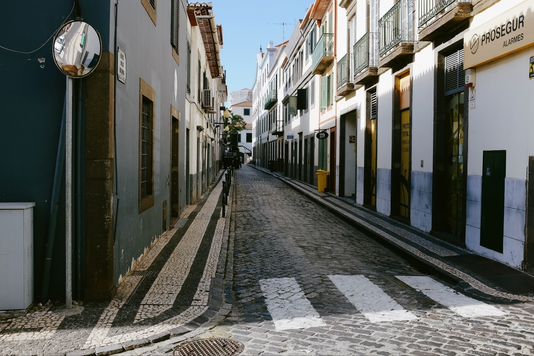 A narrow cobblestone street lined with traditional buildings, featuring balconies, shuttered windows, and decorative pavements. A convex mirror and a street sign are visible on the left, with a yellow bin on the right.