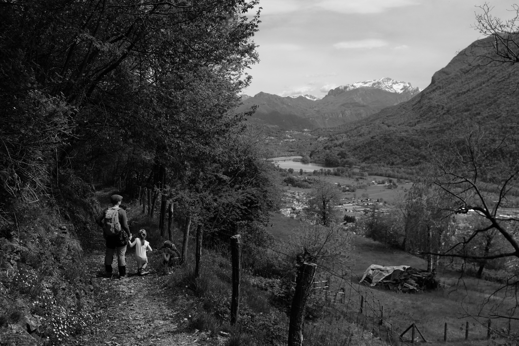 Black-and-white photo of a hiking path with a woman and two children in the foreground, and a valley sprinkled with houses and mountains covered in snow in the background