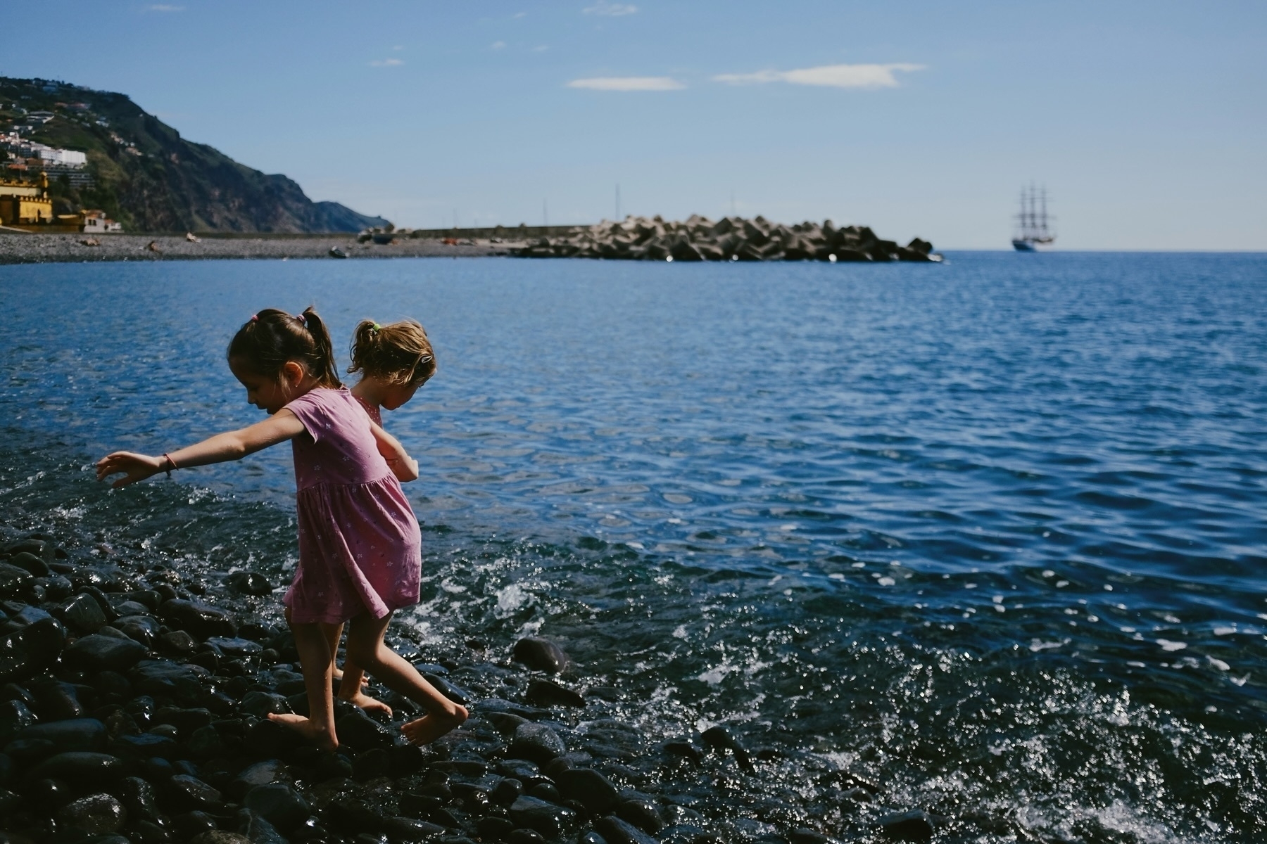 Two children in pink dresses walk on a rocky beach by the sea, with hills in the background and a ship in the distance under a clear blue sky.