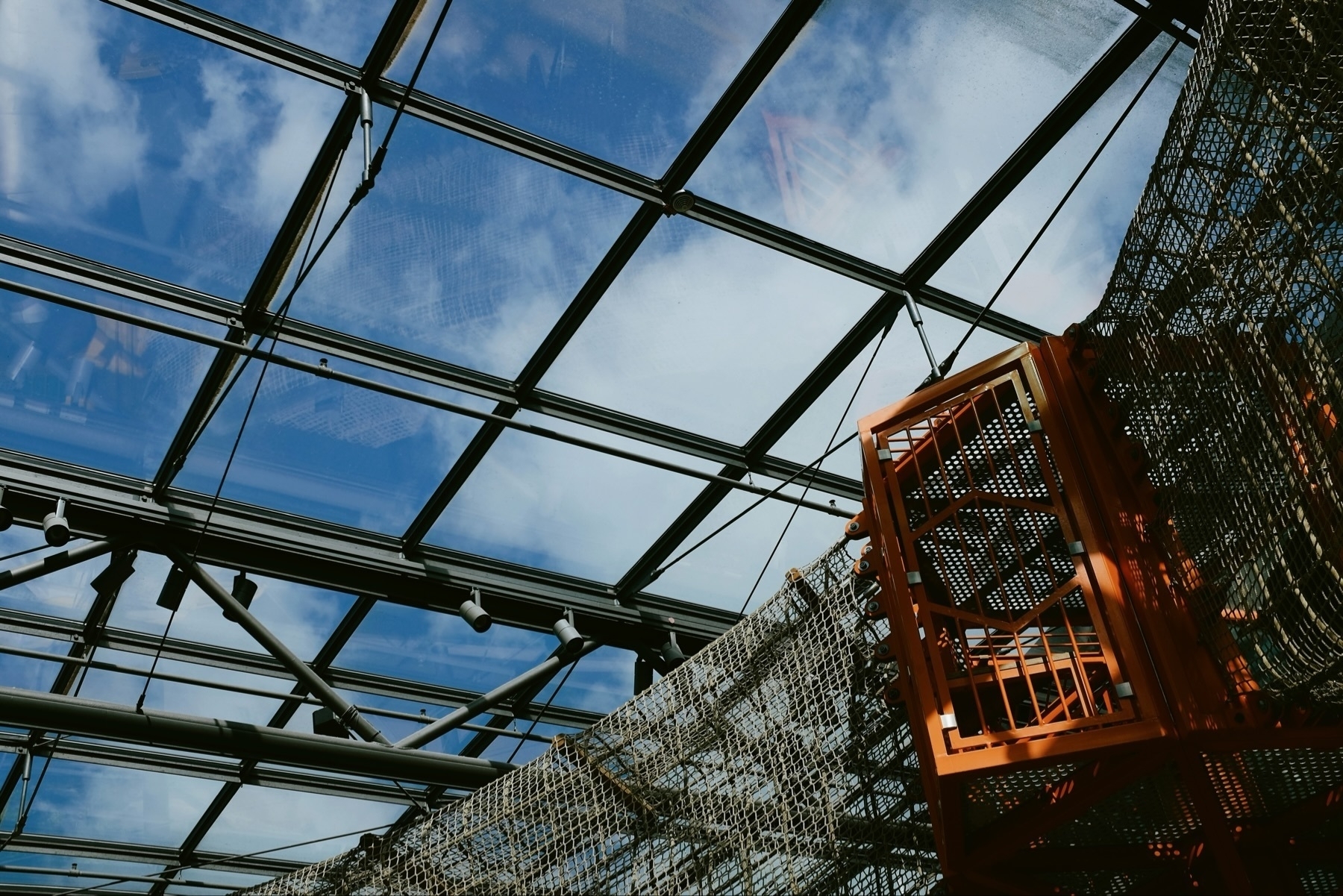 A glass roof with a metal framework, blue sky and clouds visible through it. A metal mesh structure and an orange metal cage are suspended from the ceiling.