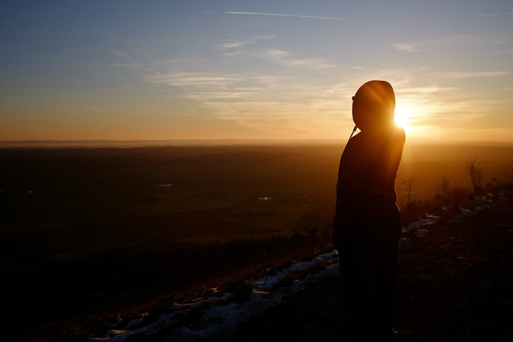 A person in a hoodie stands on a hillside gazing at the sunset.