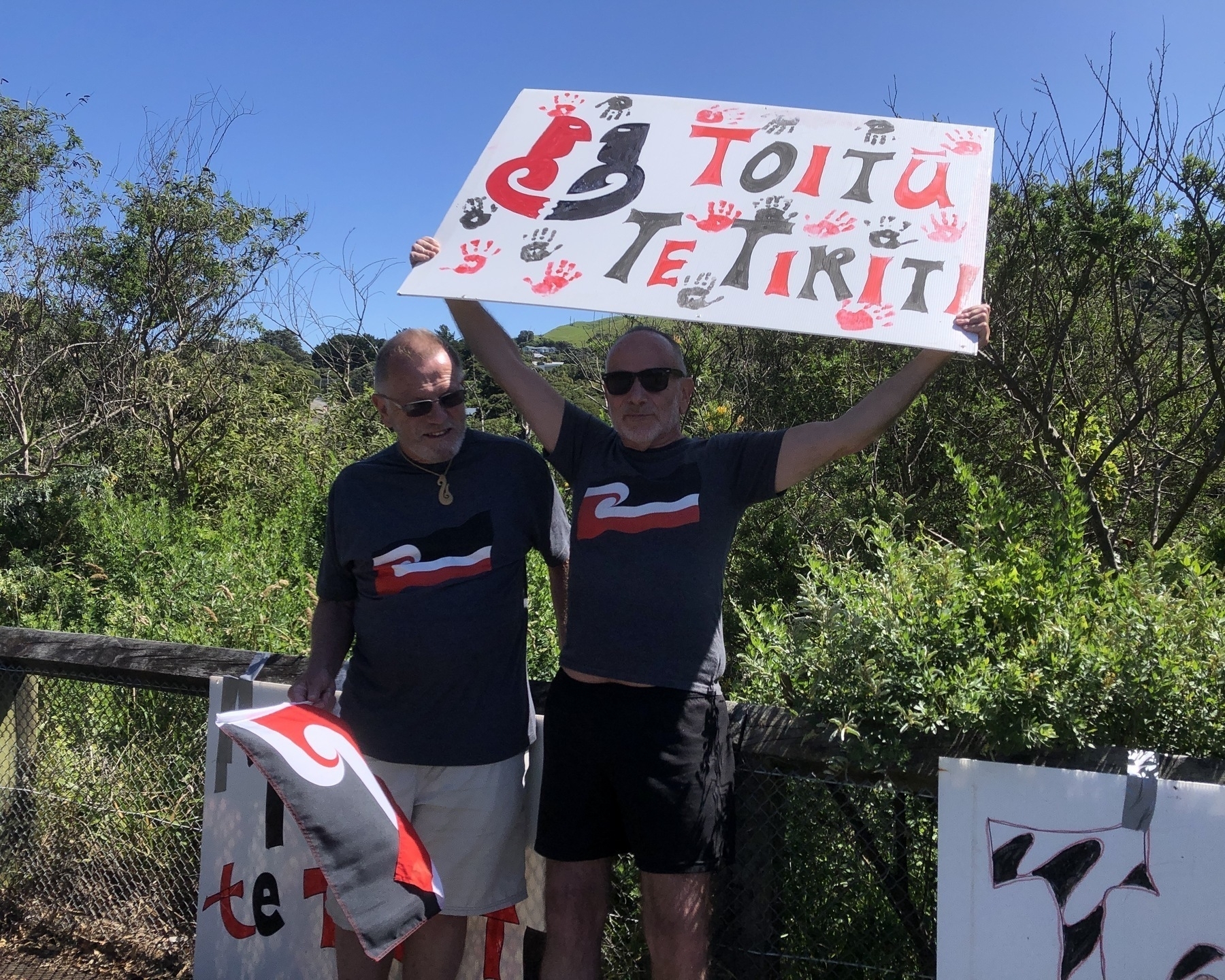 Auto-generated description: Two people are holding signs with text and designs, standing outdoors near greenery.