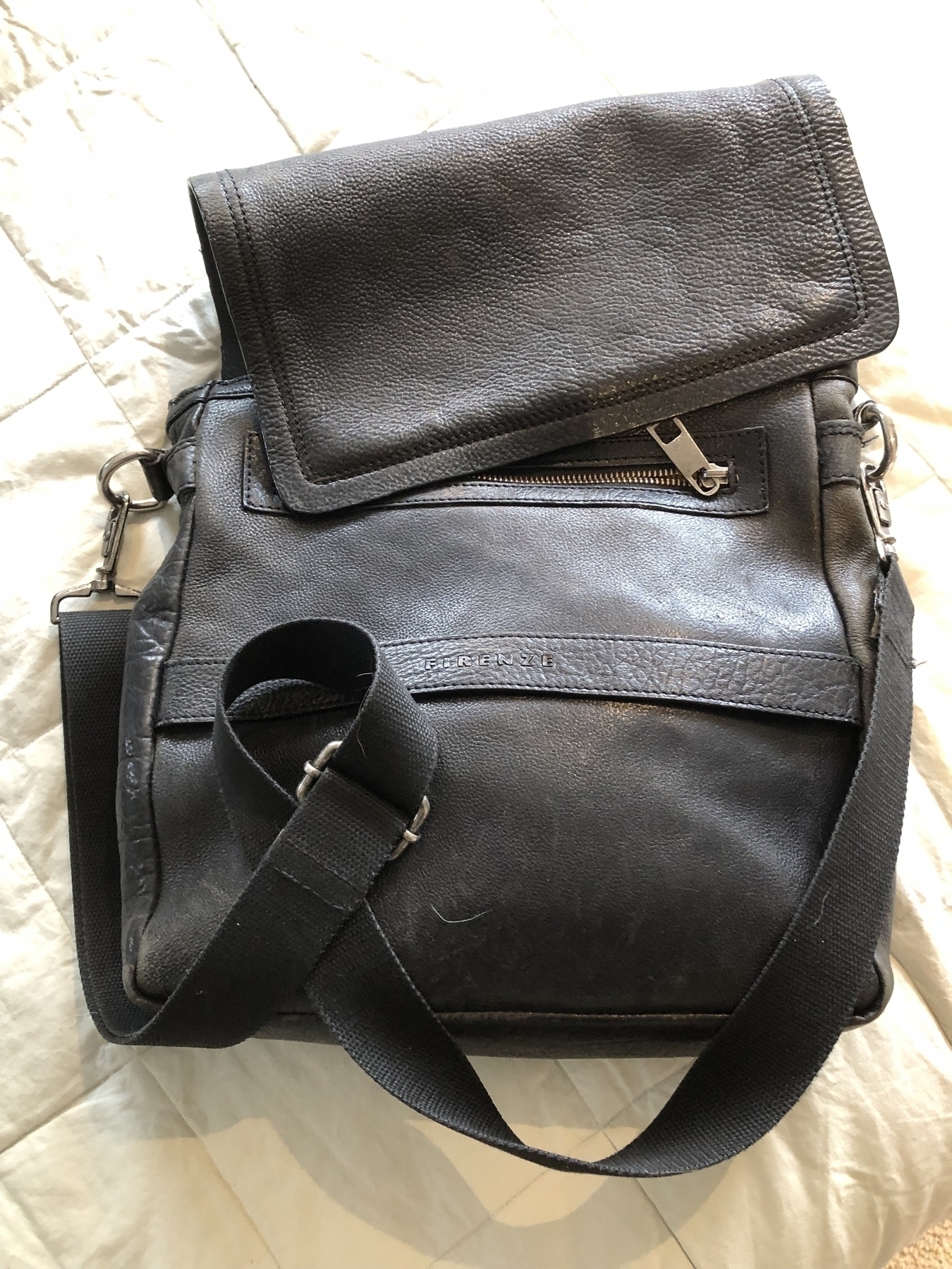 A black leather shoulder bag with a flap and adjustable strap is laid on a light-colored surface.