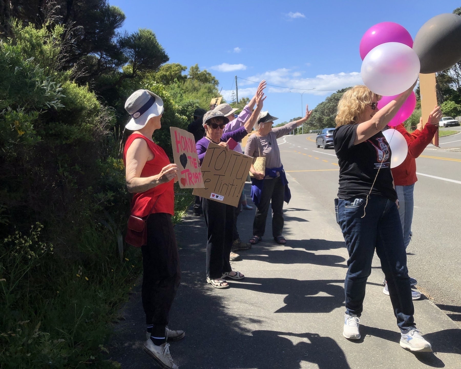 Auto-generated description: A group of people are standing on the side of a road holding signs and balloons.