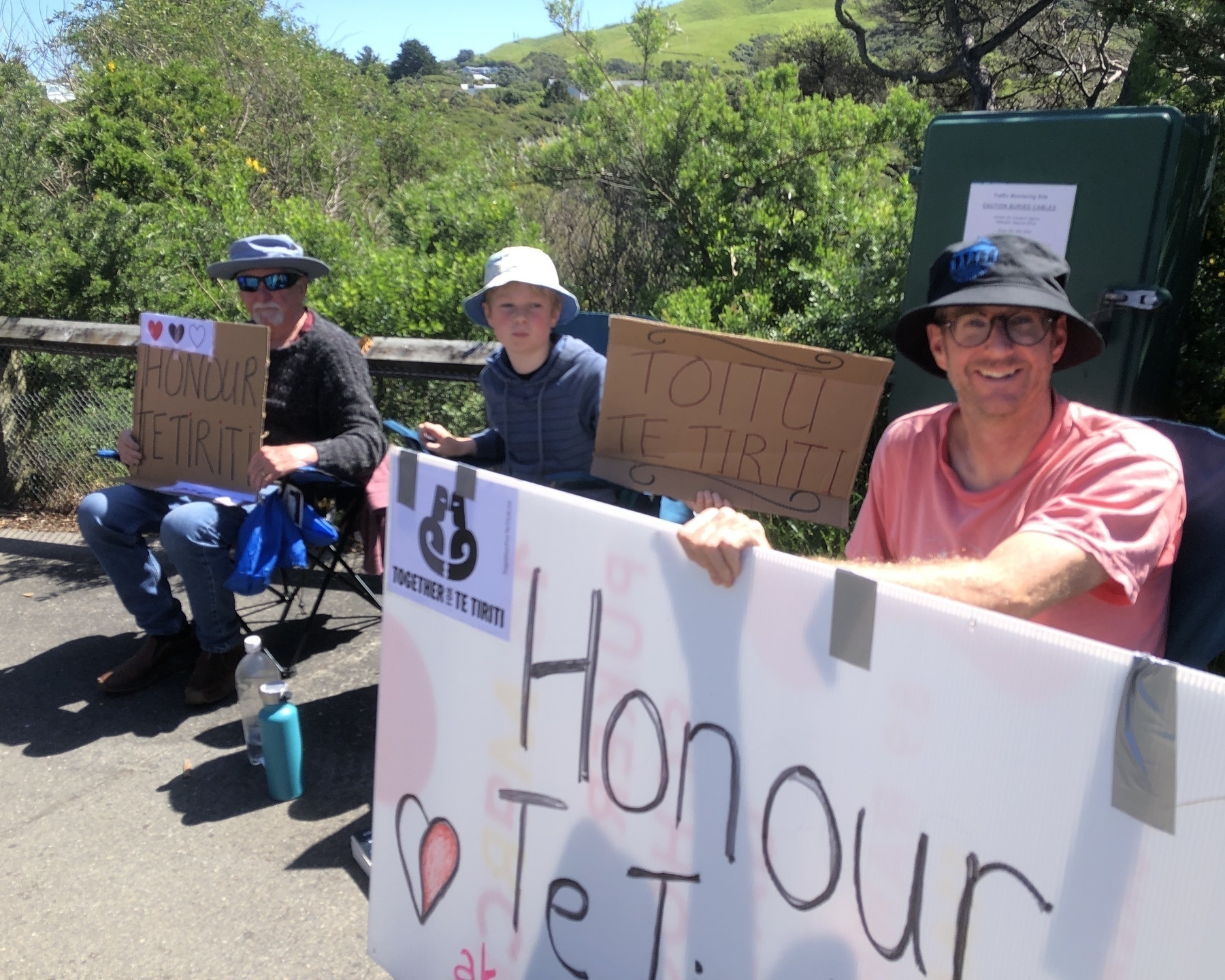 Auto-generated description: Three people holding signs reading Honour Te Tiriti and Toitu Te Tiriti are sitting outside on a sunny day.