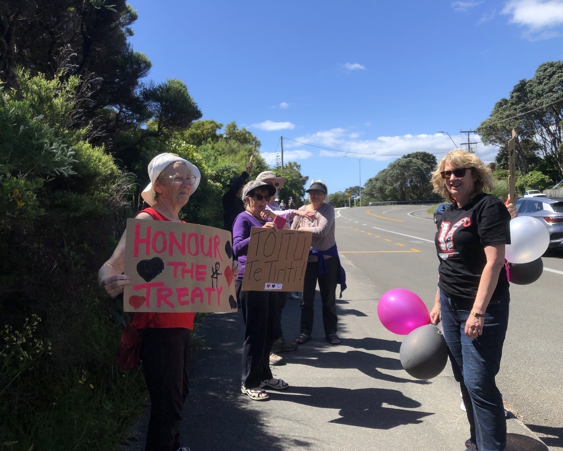 Auto-generated description: People are holding signs and balloons along the side of a road under a clear sky.