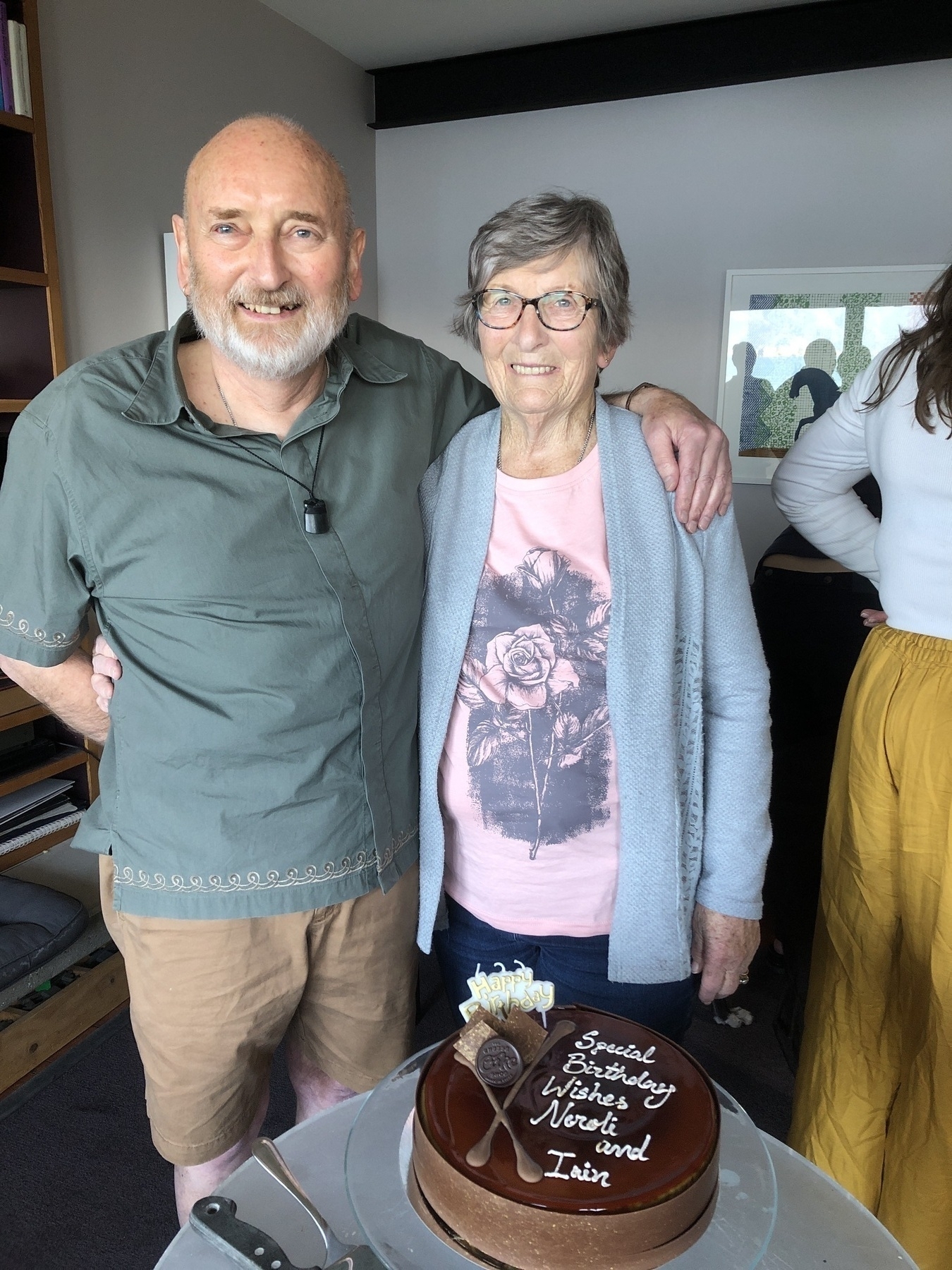 A smiling elderly couple stands next to a chocolate cake with a birthday message.