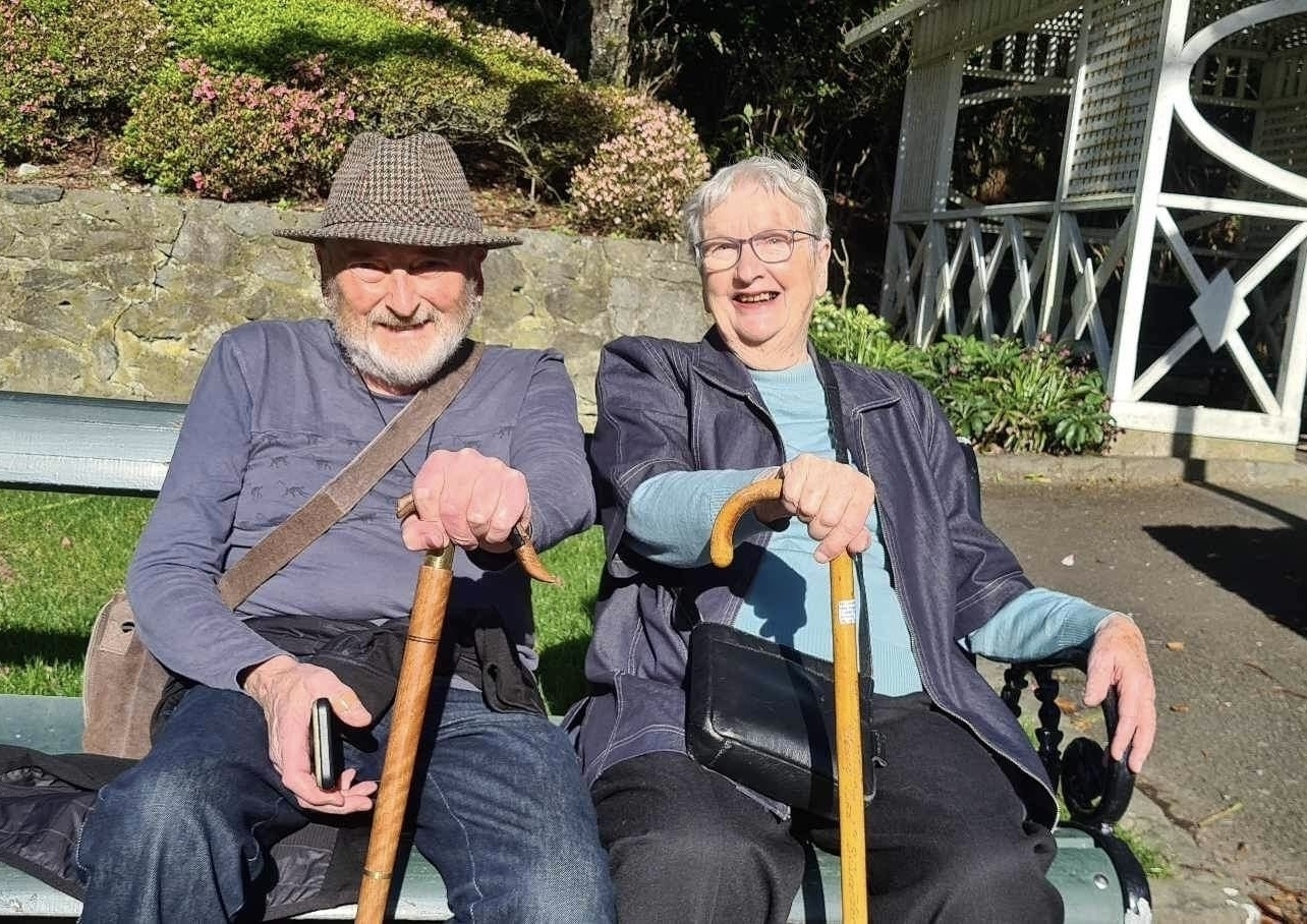 Grey-haired man and woman sitting on park bench showing their almost matching walking sticks