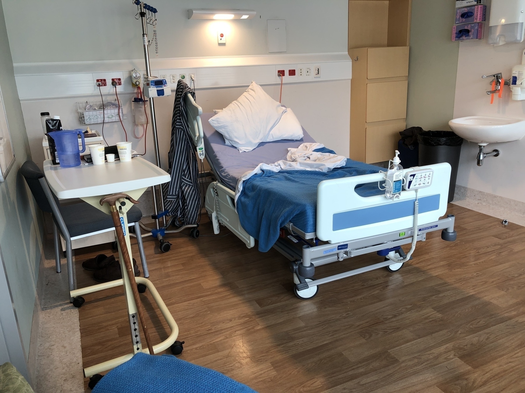 Auto-generated description: A hospital room features an unoccupied bed, a bedside table with personal items, medical equipment, and a sink area.