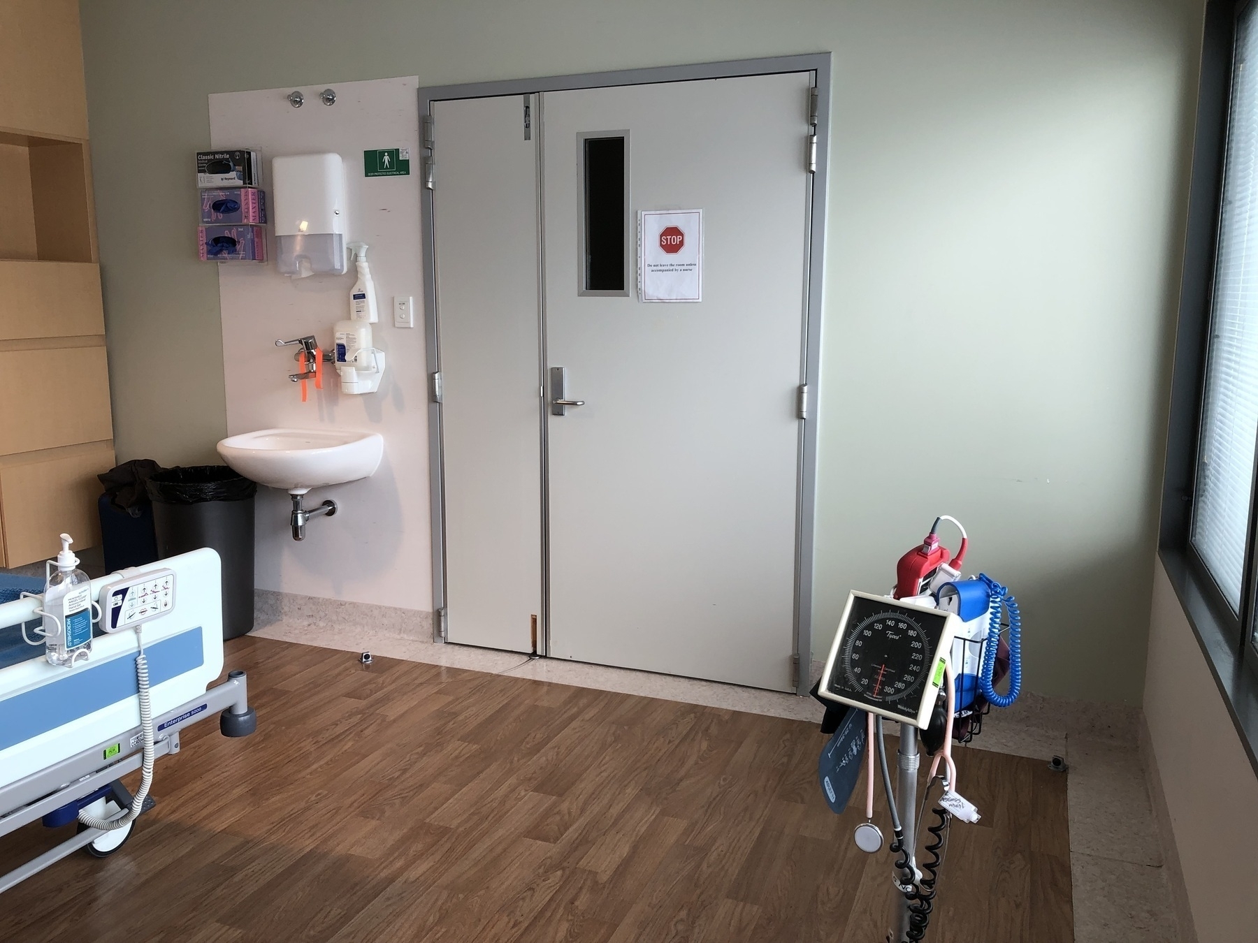 Auto-generated description: A hospital room features a medical bed, a handwashing station, medical equipment, and a closed door.