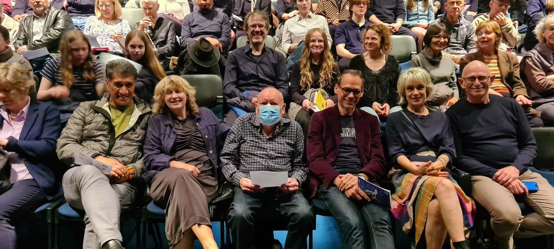 Auto-generated description: A large group of people is seated in an auditorium, with some individuals in the front row smiling for the camera.