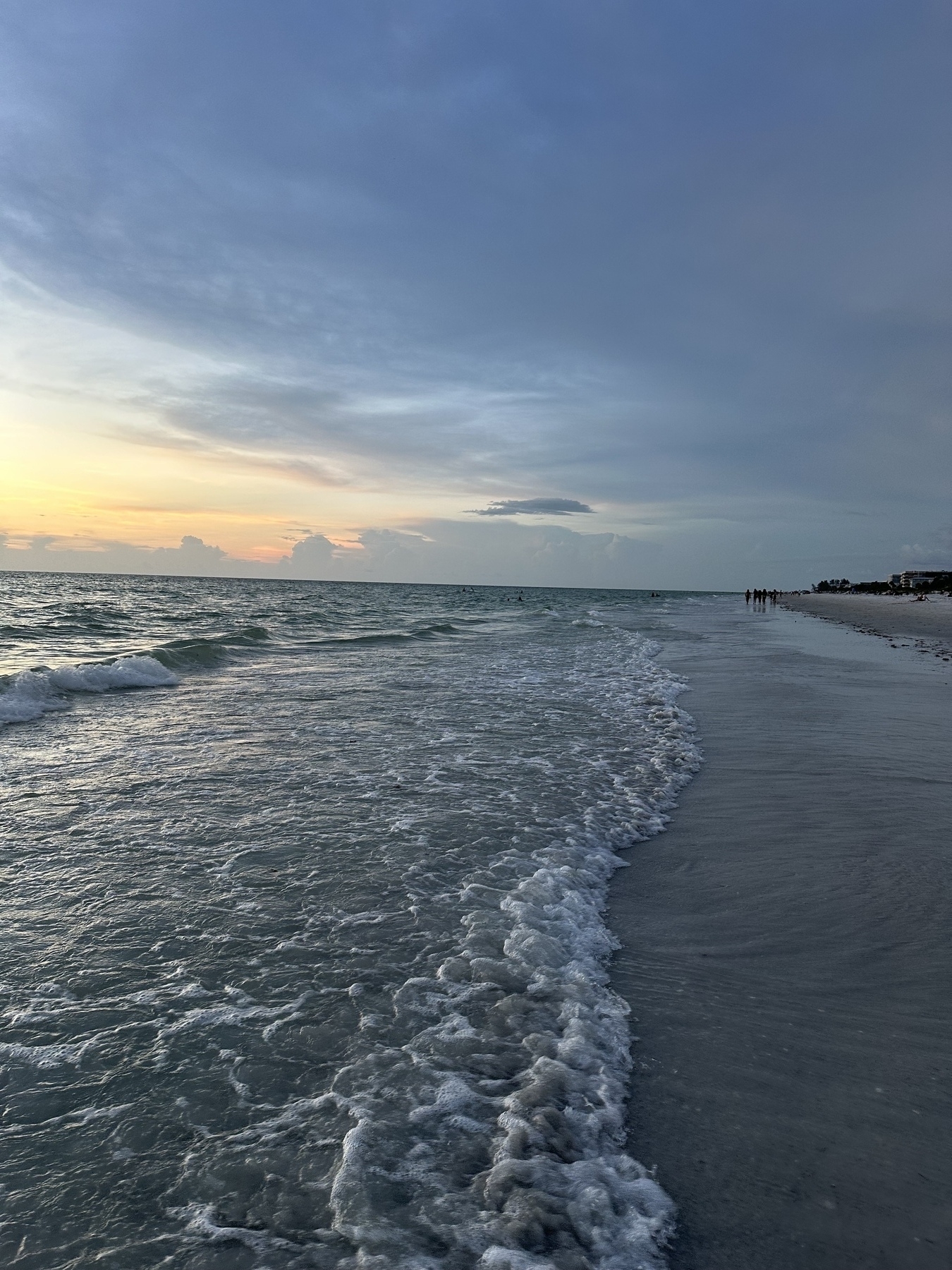 Gentle waves wash ashore on a sandy beach under a cloudy sky at sunset.