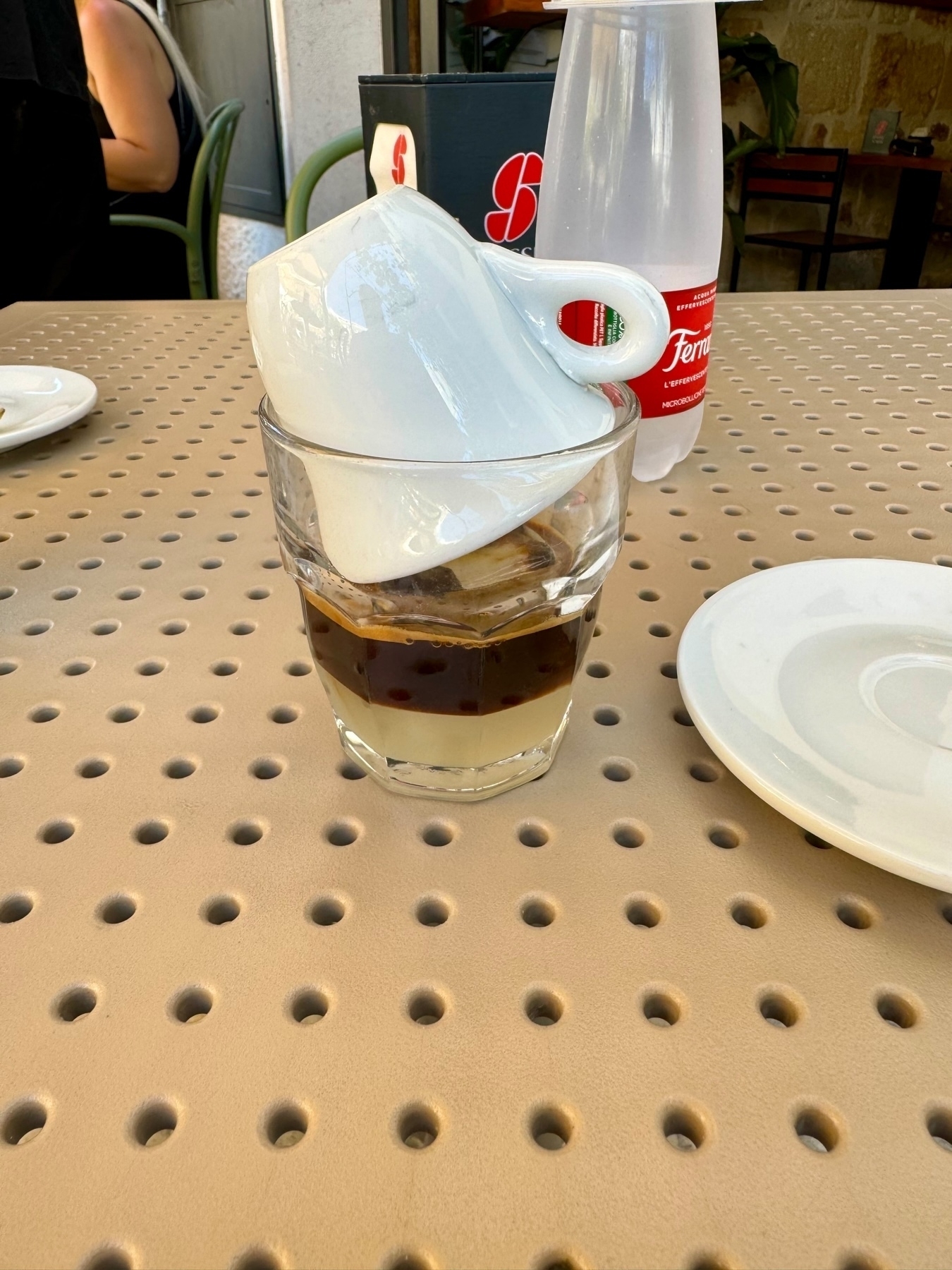 A white coffee cup is placed upside down inside a glass filled with a two-layered liquid, coffee and almond syrup. The setup is on a perforated beige table next to an empty white plate and a bottle of Ferrandina water.