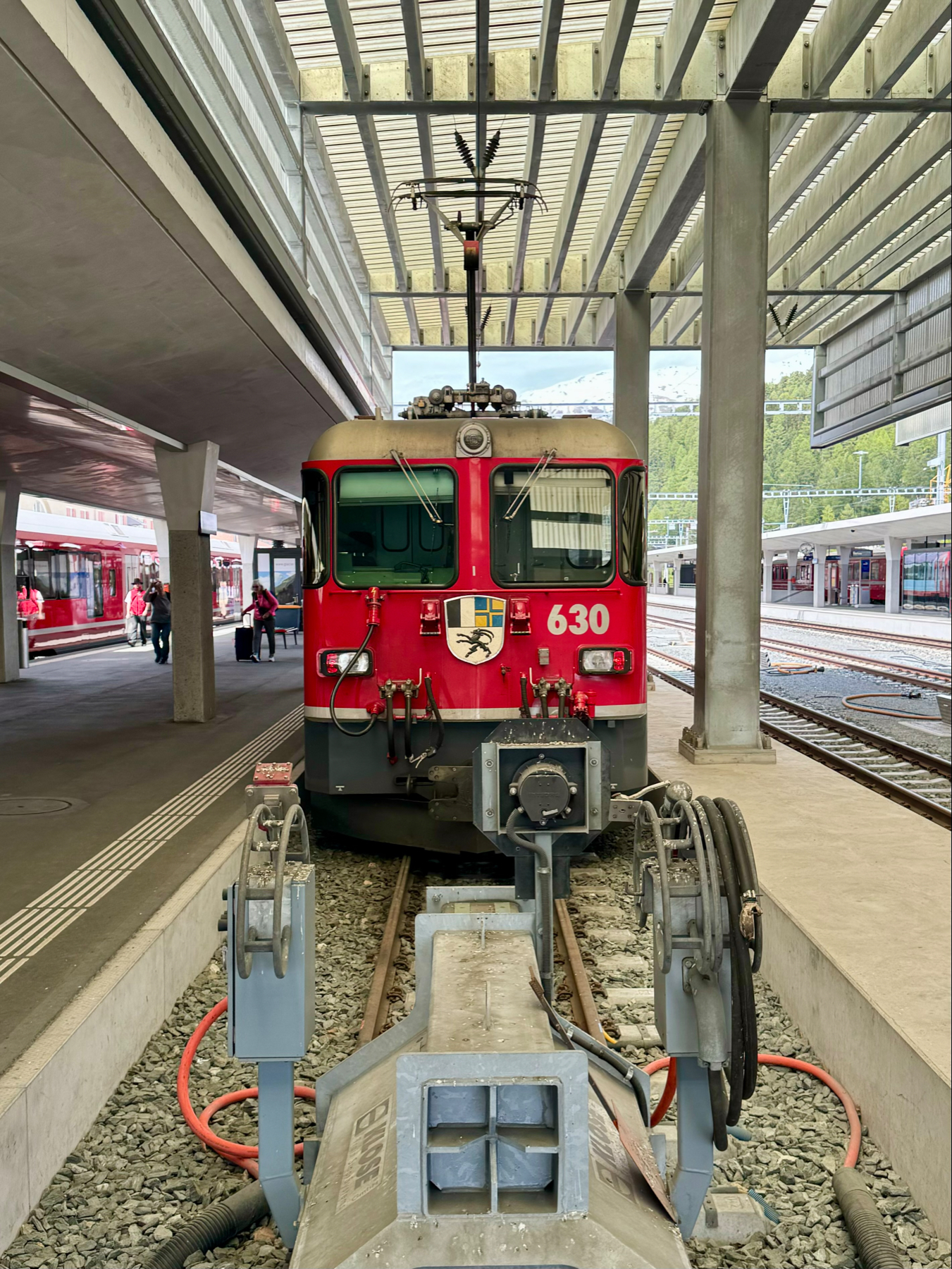 A red and white electric train at a platform with overhead power lines, a pantograph raised to connect with the electric supply, and other trains in the background.
