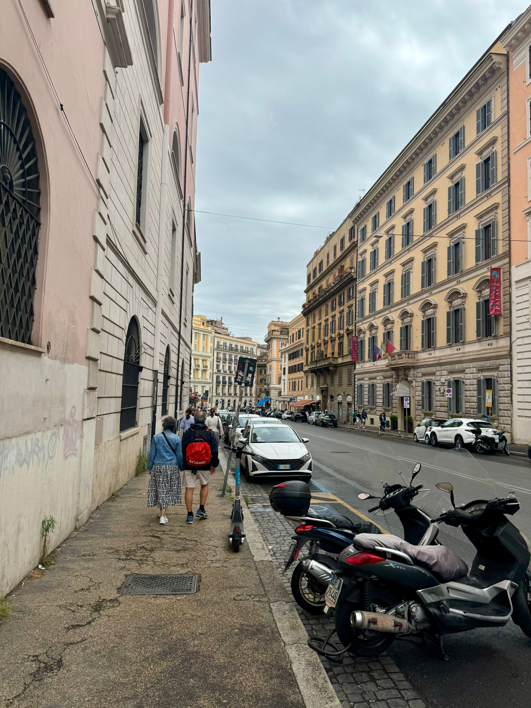 A street scene in Rome. The view shows historical buildings with classic architecture on both sides. Scooters and motorcycles are parked along the cobblestone curb. People walk on the sidewalk under an overcast sky.